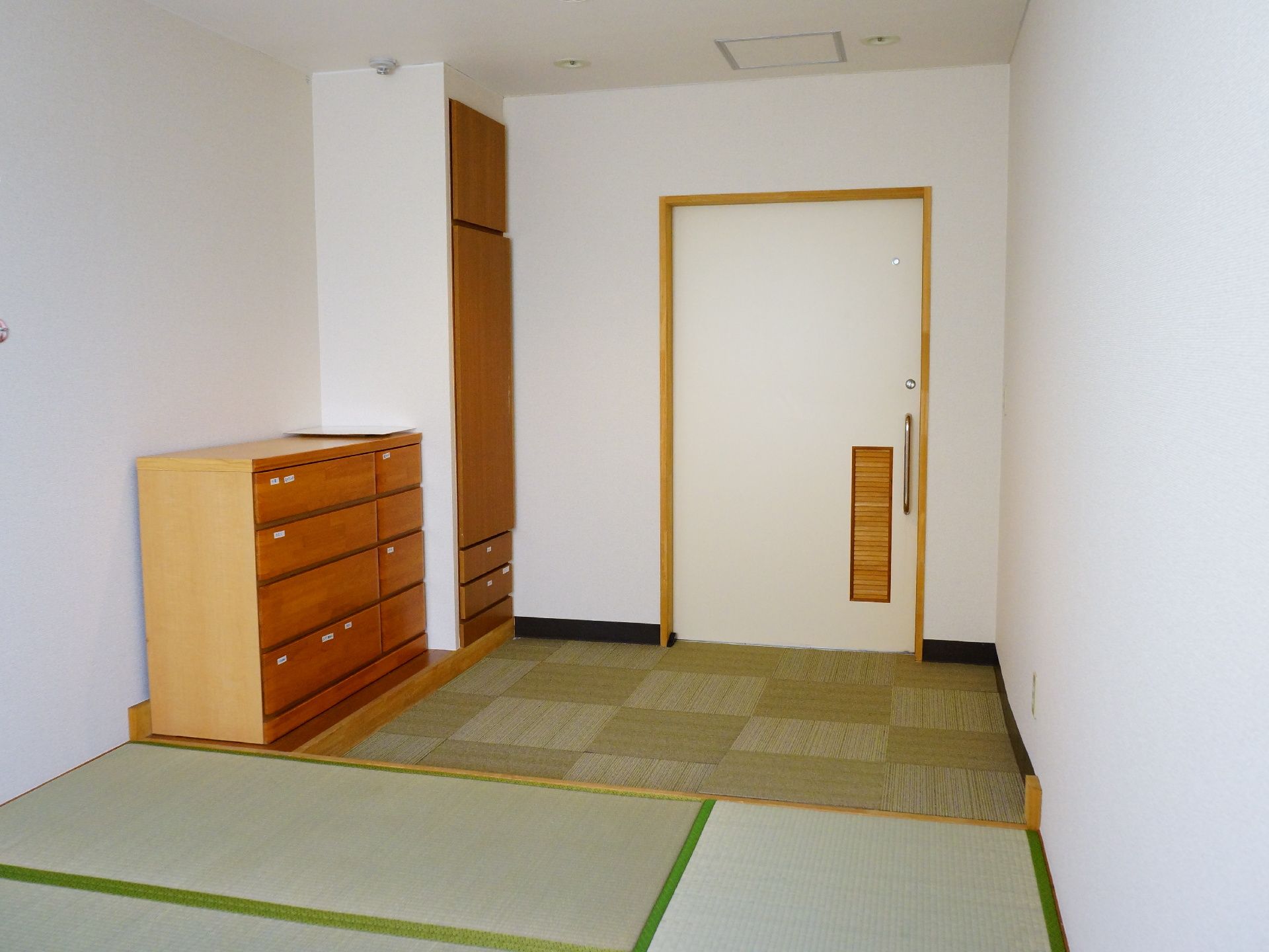 正面奥には扉がある。左側には収納スペースがあり、箪笥がその隣に置かれている。壁と天井は白色に塗られている。