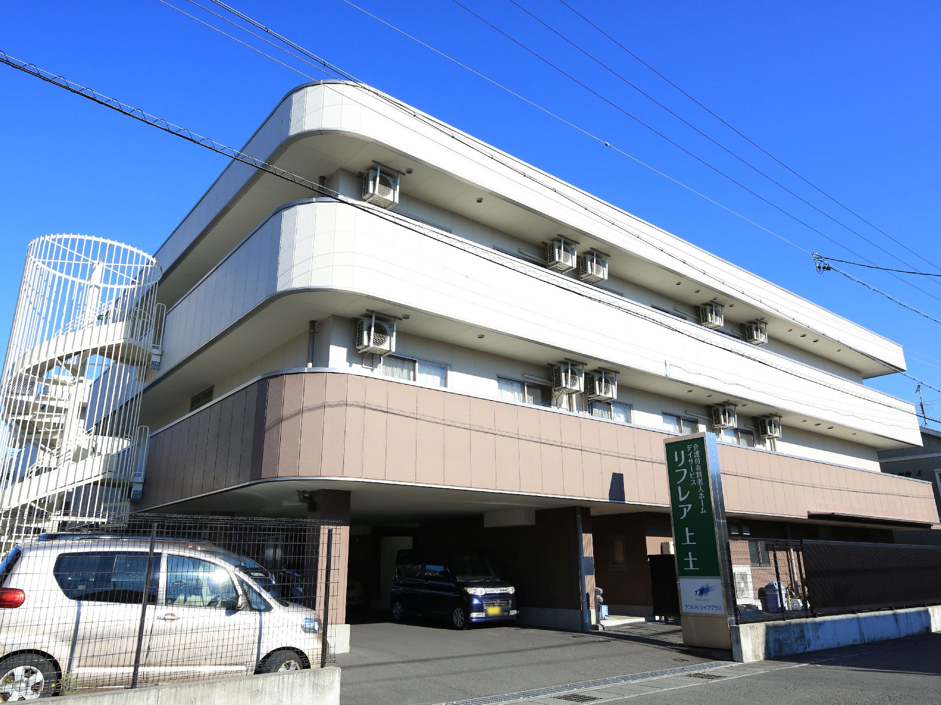 ３階建ての大きな建物がある。外側にはベランダが設置されている。建物の周囲はフェンスで囲まれ、入り口に大きな看板が掛けられている。