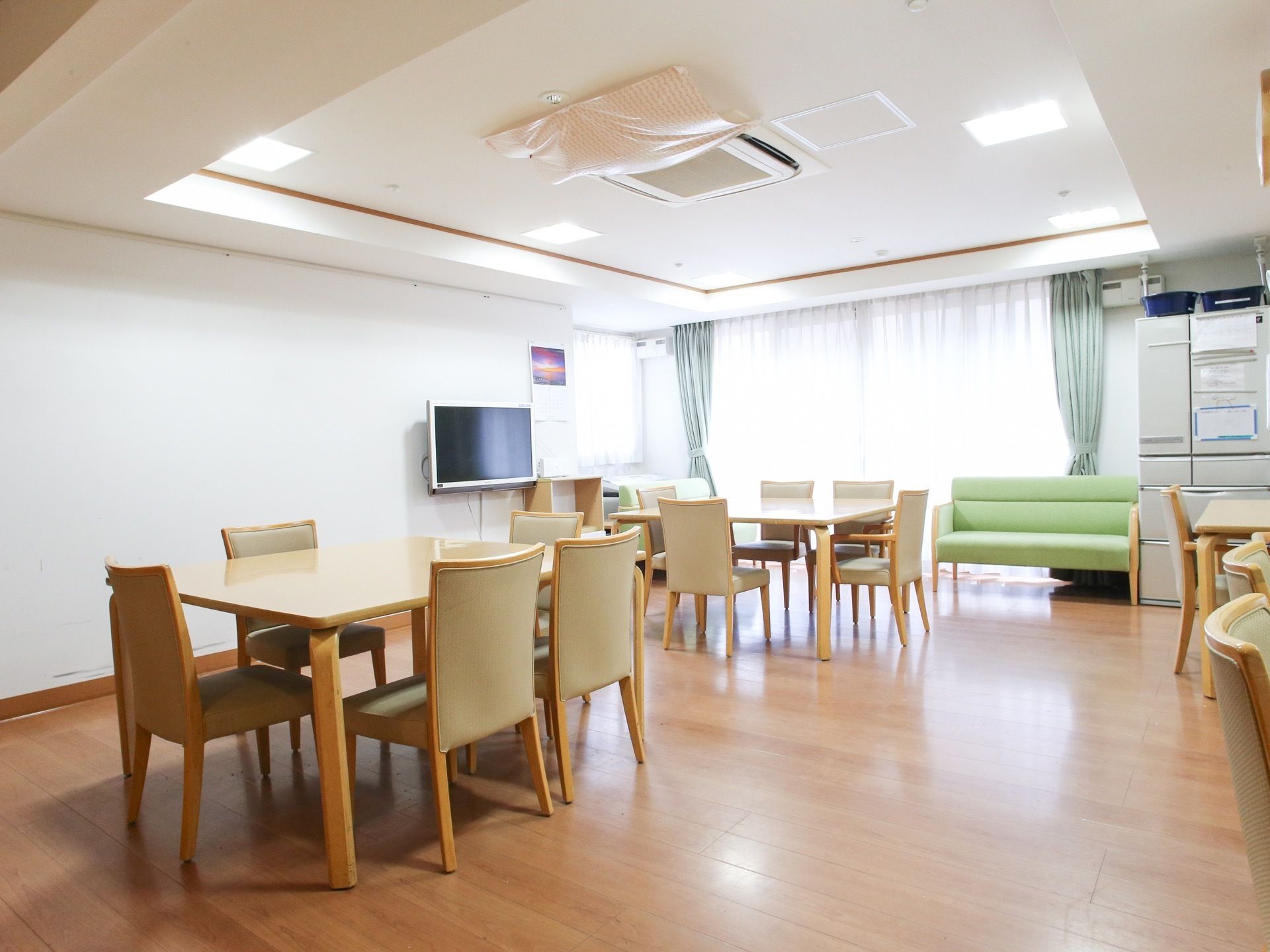 ５人がけのテーブルセットが配置された食堂。奥には大型の冷蔵庫とソファが置かれ、背後には大きな窓がある。左の壁にはテレビが設置されている。