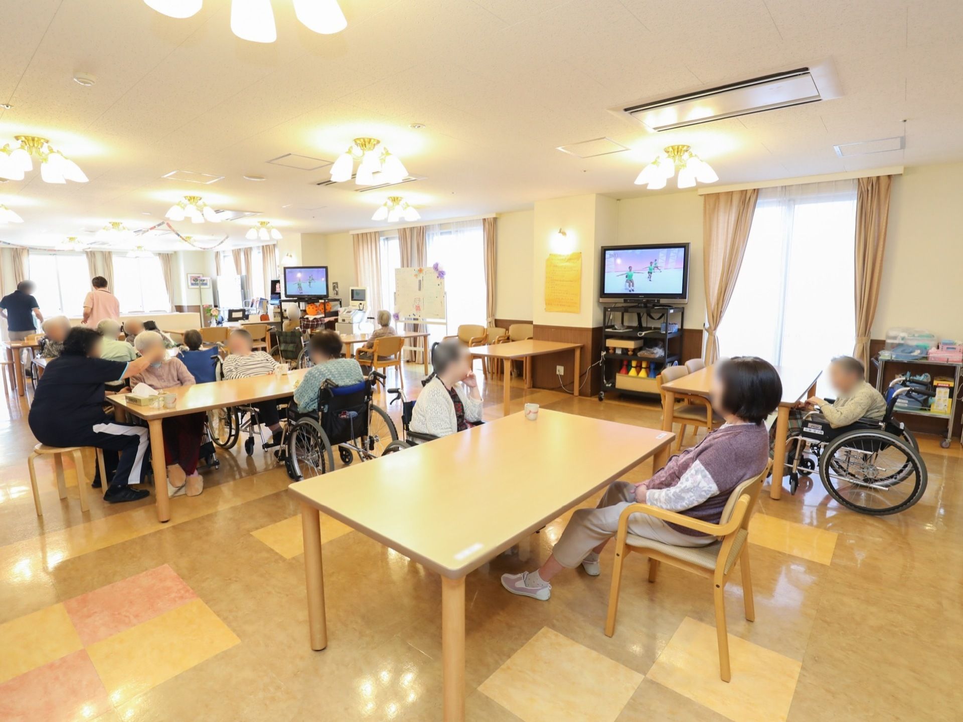 入居者様がお茶を飲まれている。窓側にテレビが2台設置されている。体操番組が流れており、テレビの前で体操をされている入居者様もいる。