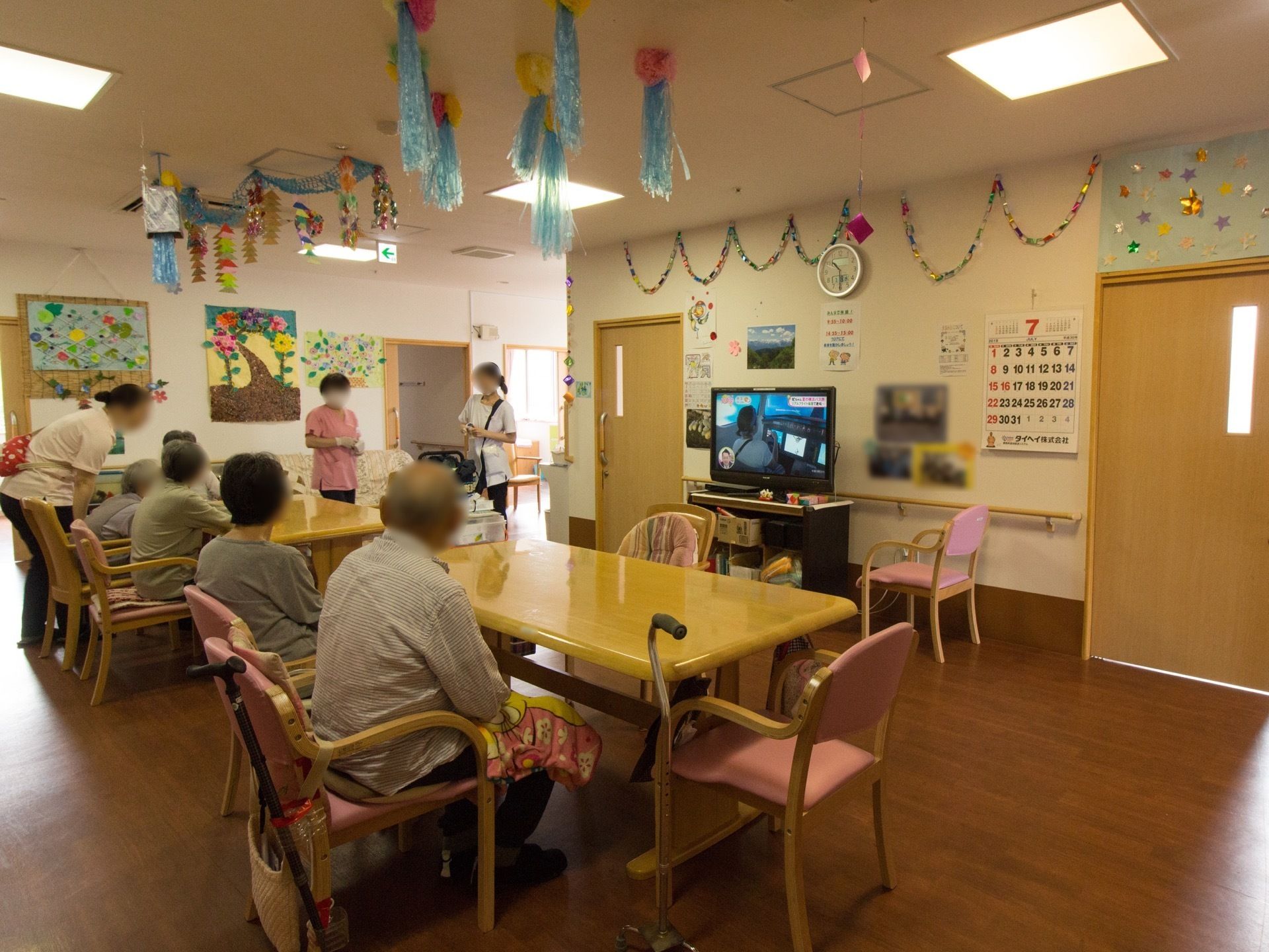 ピンクの椅子が並べられたテーブルセットが置かれている。壁には時計やカレンダーが貼られ、液晶テレビが用意されている。