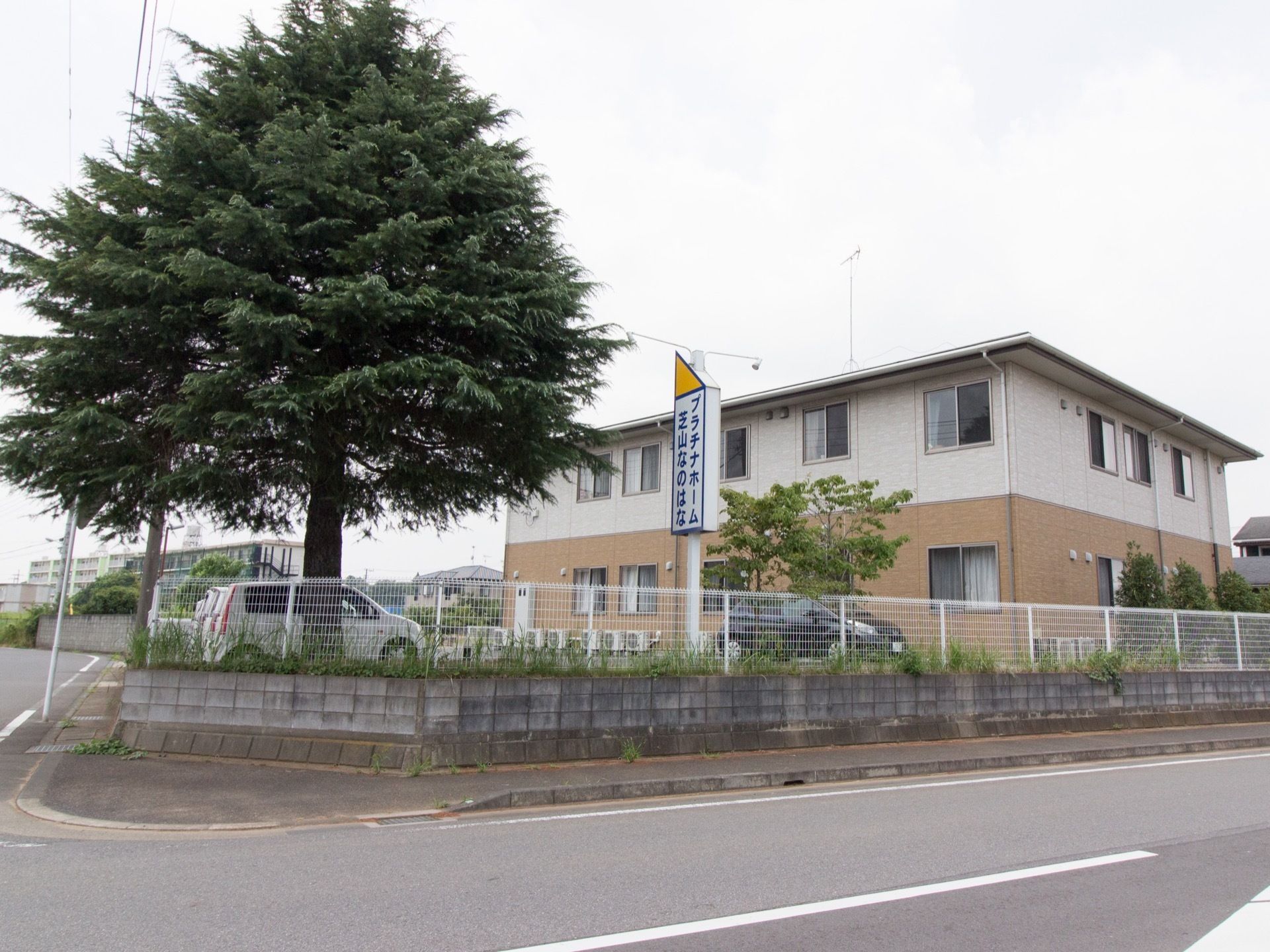 施設の敷地に看板を設置している。遠くからも見える大きな樹木が生えている。施設前は駐車スペースが広がっている。