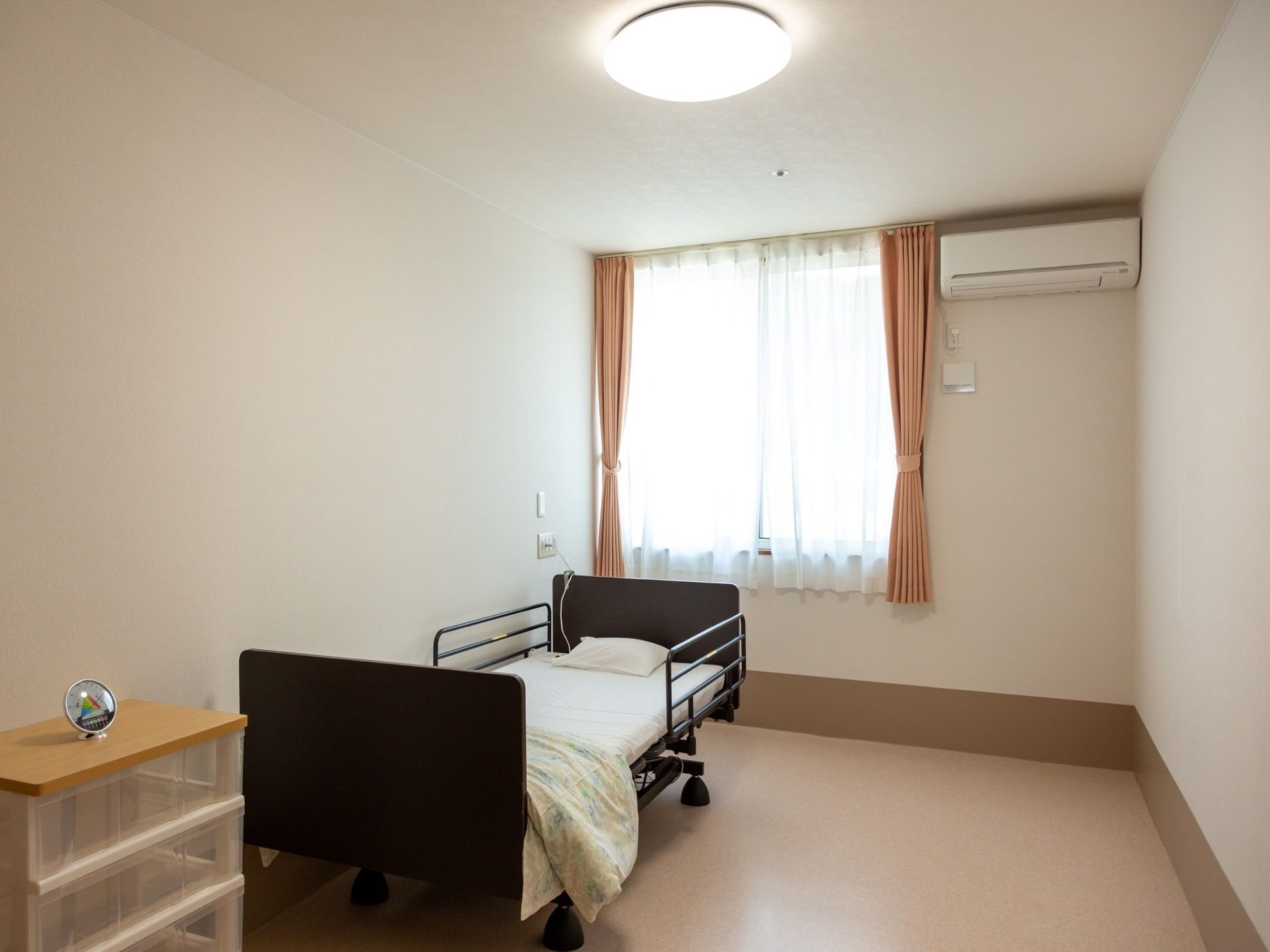 居室の壁際に介護用ベッドが置かれている。窓にはオレンジのカーテンとレースカーテンが下がっている。エアコンが完備されている。
