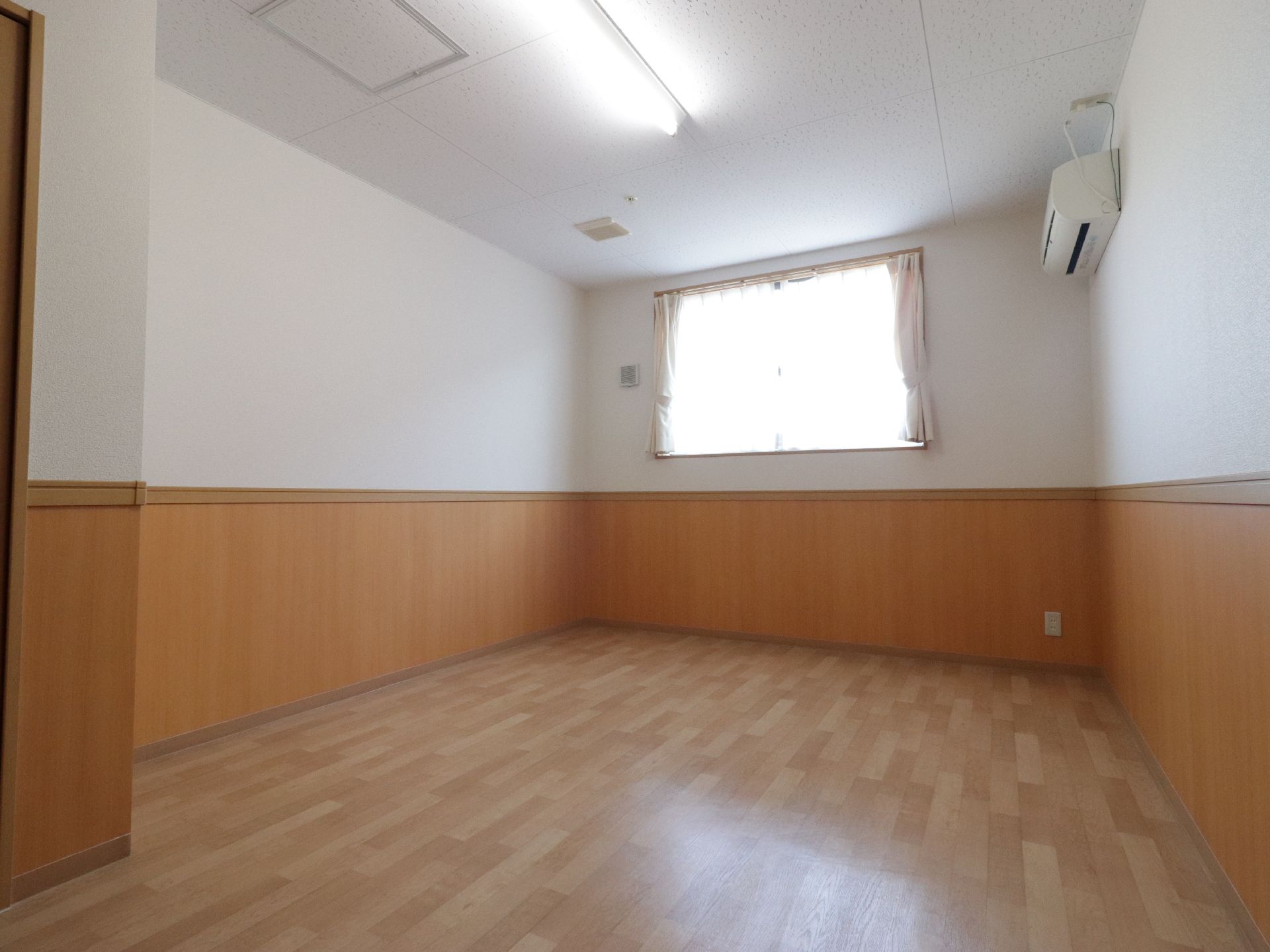 居室の奥には腰高窓があり、白いレースとアイボリーの厚地カーテンが掛けてある。左側の壁には、エアコンが設置されている。