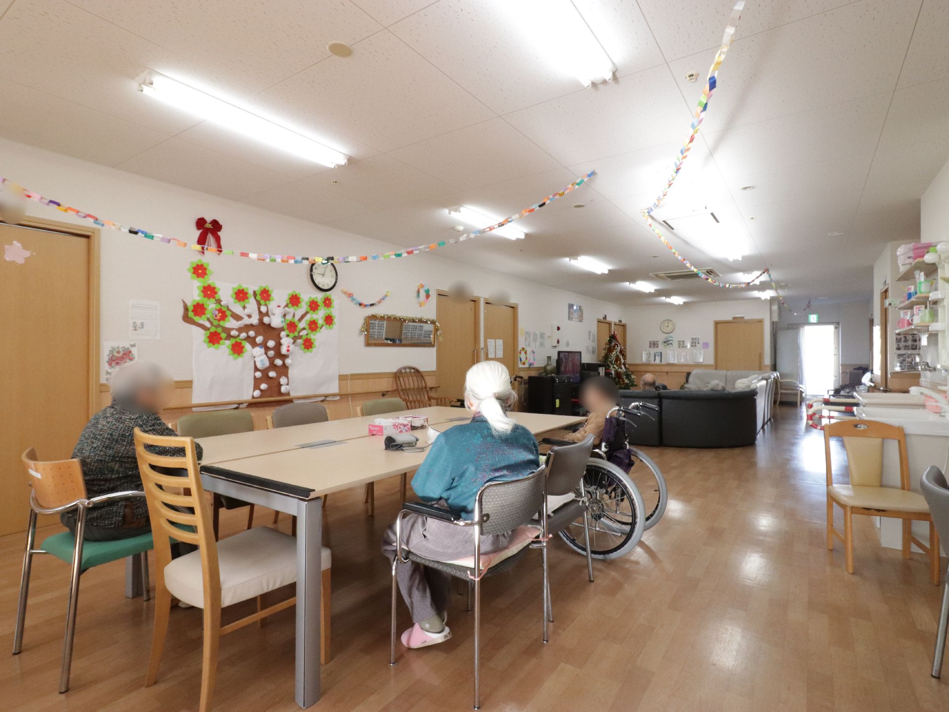 ライトブラウンの大きなテーブルの周りに、３名の入居者様が座っている。奥の方には、クリスマスツリーが飾ってあるのが見える。