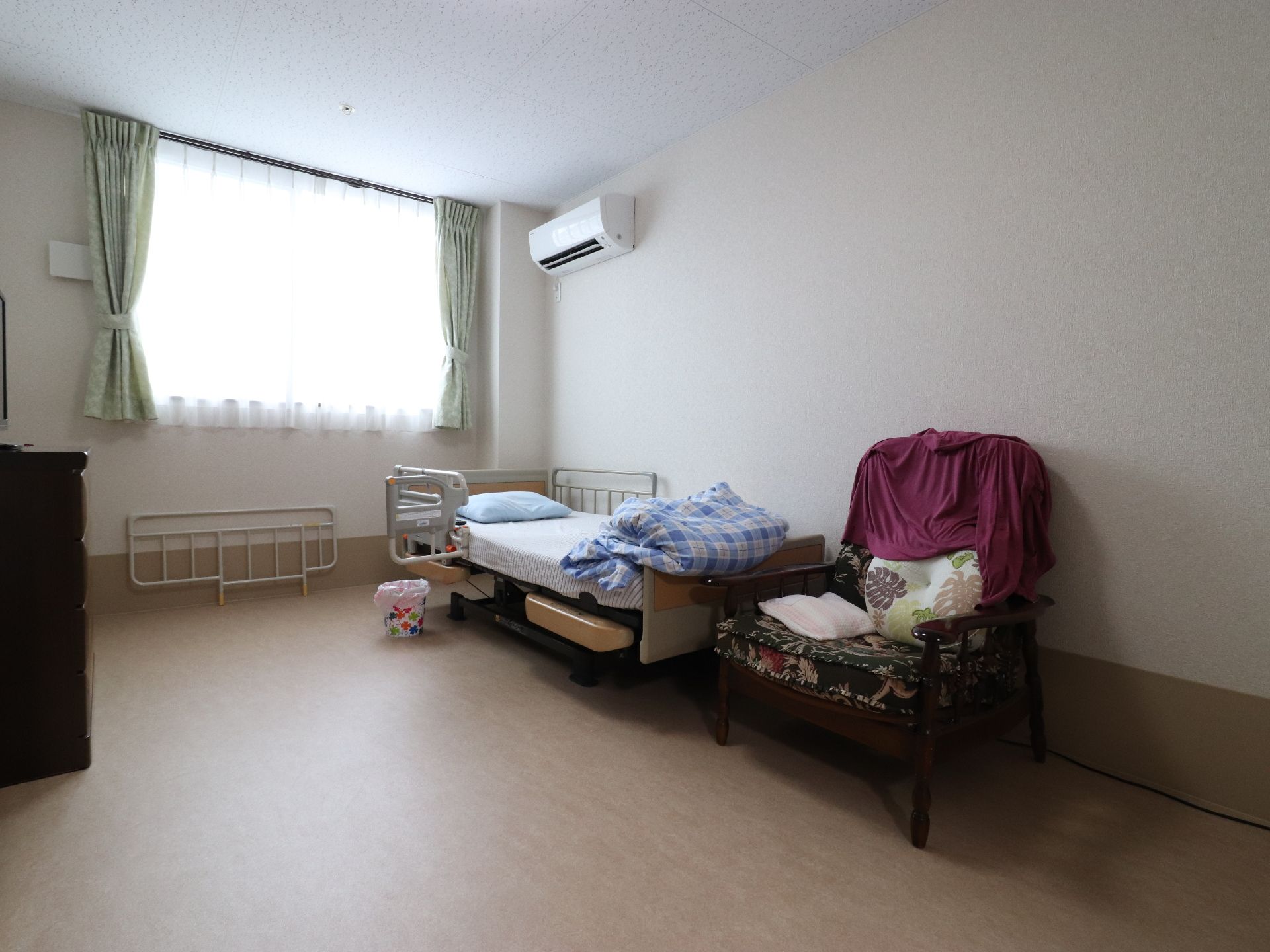 居室には大きな腰高窓があり、日差しが射し込んでいる。右側の壁にはエアコンがあり、その下にはフェンスが設置されたベッドがある。
