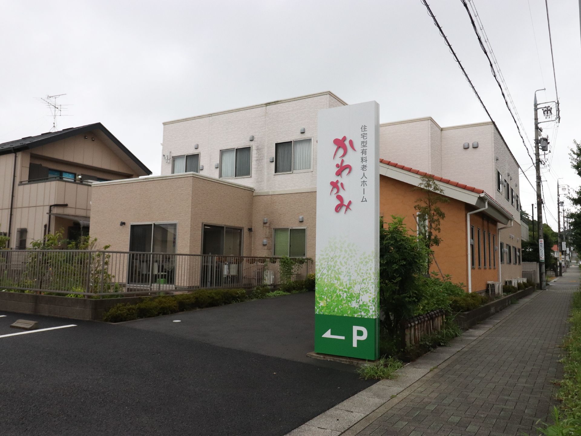 施設の入り口前には様々な植物が植えられている。その前面には白地に赤い文字で施設名が書かれた看板が設置されている。