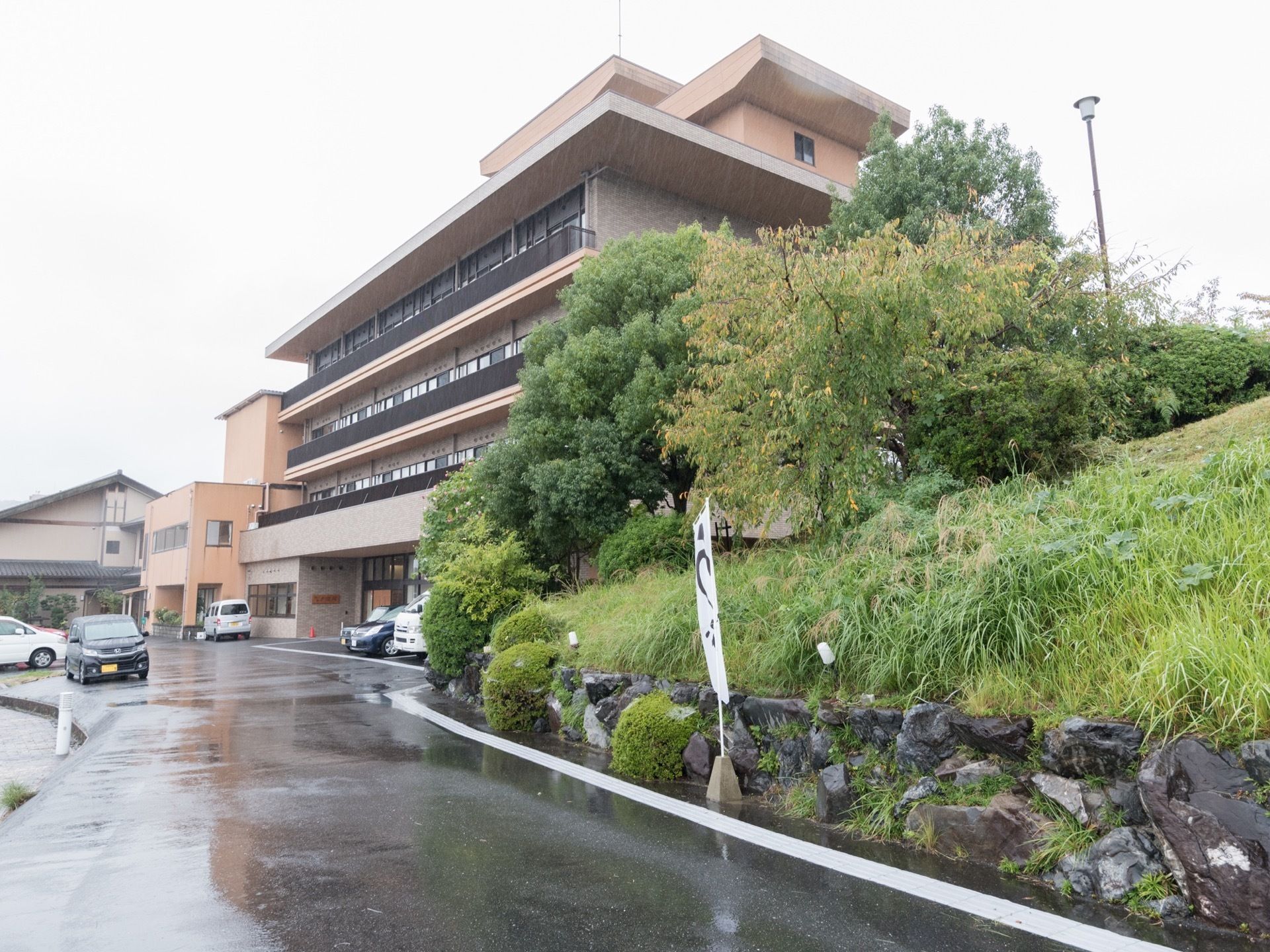 5階建ての建物で、施設までの通路の右側には石垣があり、上には草木が生えている。建物の正面に駐車場があり、数台の車が止められている。