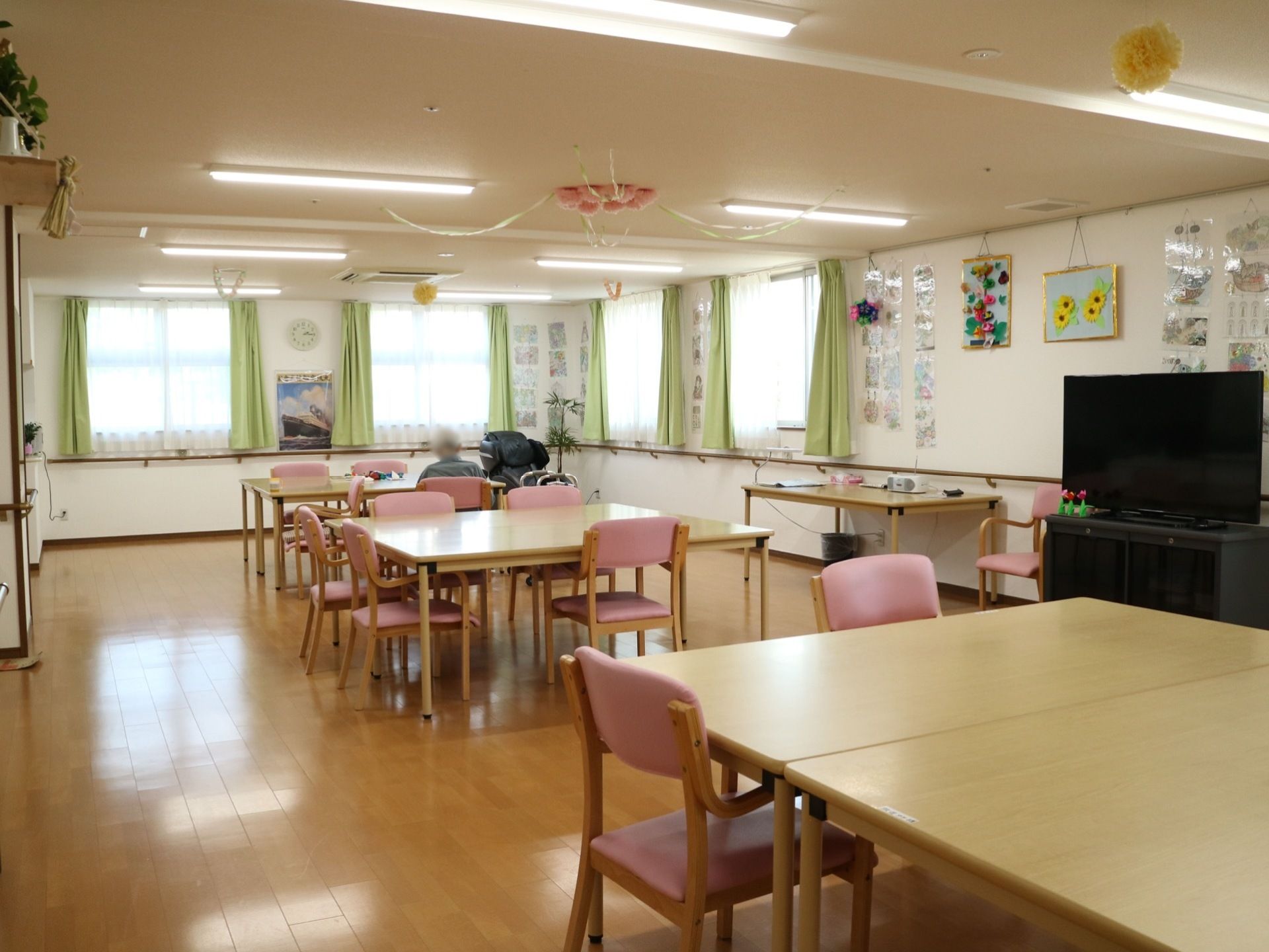 木目調のダイニングテーブルにピンクの椅子が置かれている。天井や壁に花をモチーフとした作品や、写真などが飾られている。