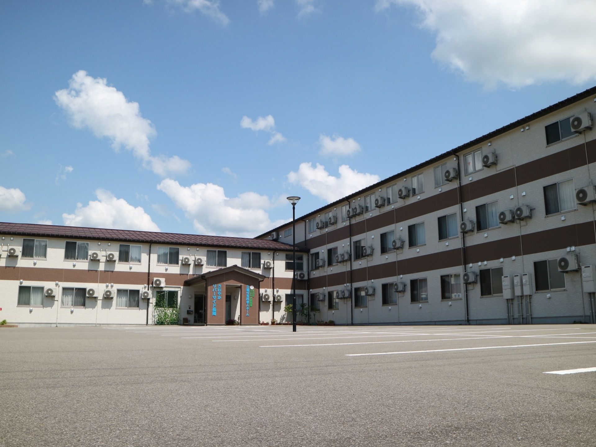 広々とした敷地に、白と茶色を基調とした３階建ての施設が建てられている。駐車スペースに外灯が設置されている。