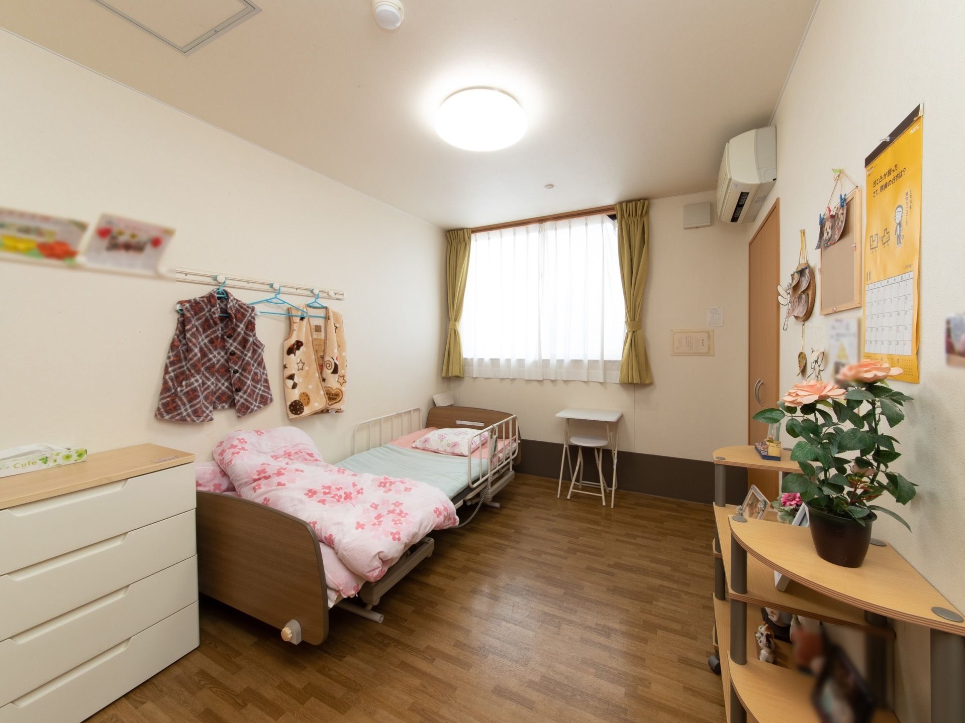 白を基調とした明るく広い居室である。介護用ベッドやエアコン、チェスト、飾り棚、カーテンが完備されている。