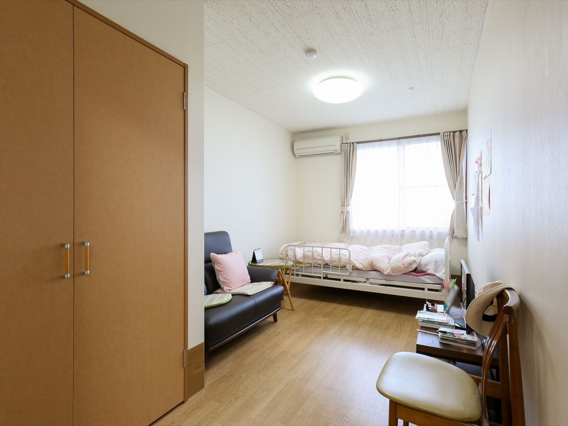 廊下側から居室内を見た図である。奥に窓があり、その前に柵のついた介護用ベッドが置かれ、布団が敷かれている。