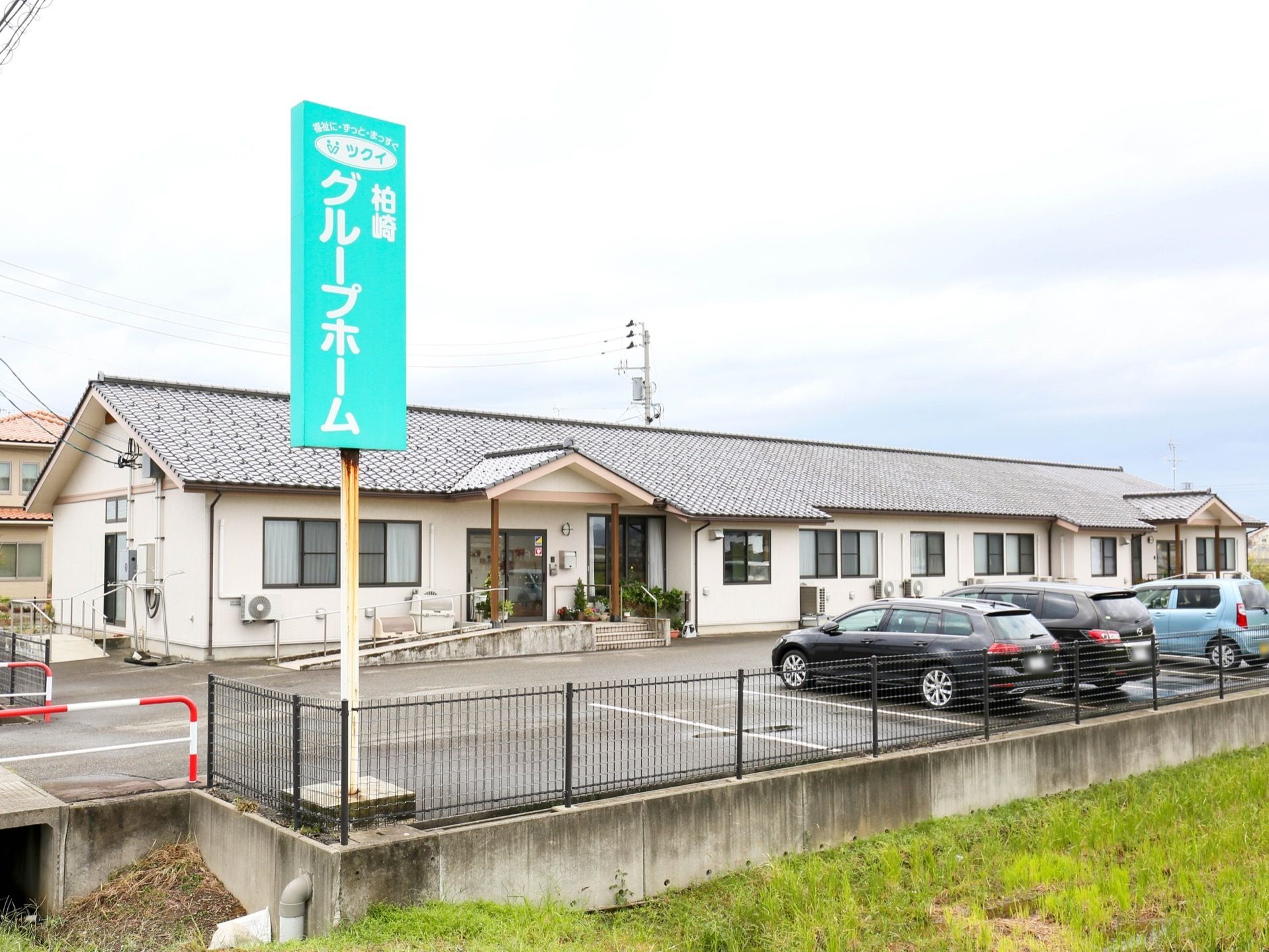 平屋建ての施設。施設の周囲が黒い柵で囲まれている。駐車スペースの脇に水色の施設名が書かれた看板が設置されている。