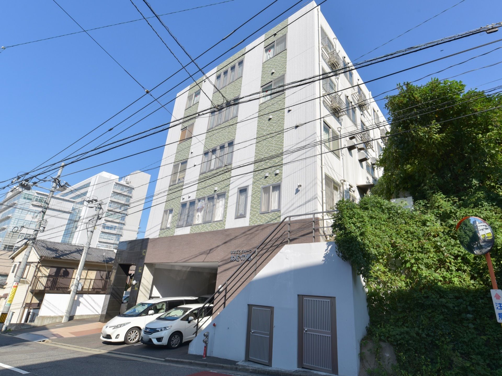 道路に面した建物の壁面には、施設名が表示されている。自動車が数台停車している。付近には自然の緑が生い茂っている。