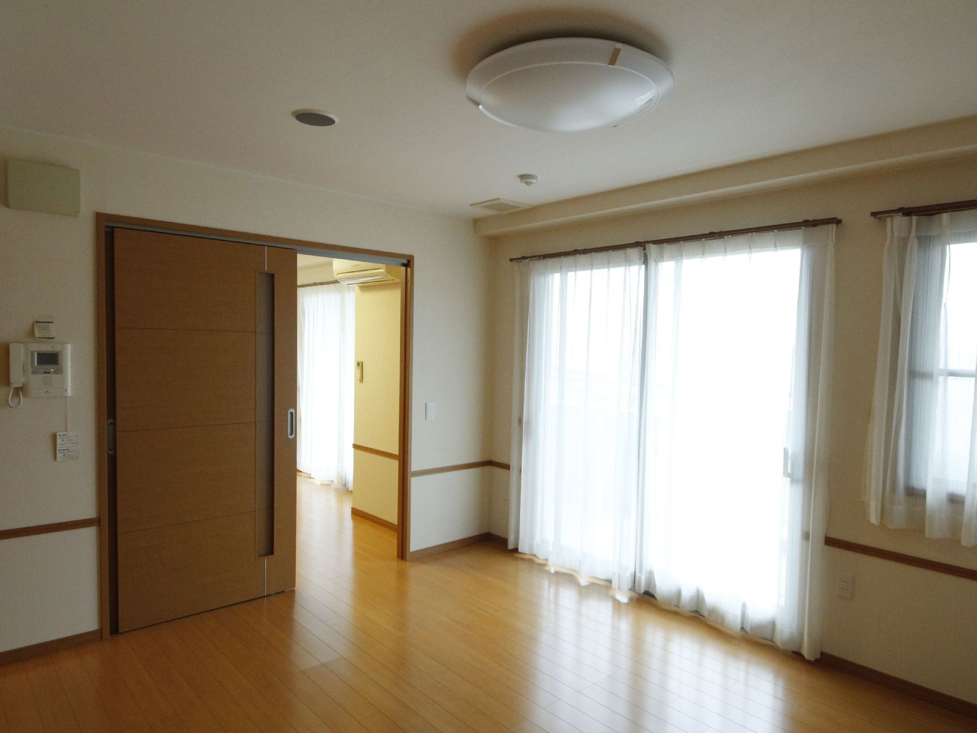 居室の床は全面フラットなフローリングである。複数設けた大きな窓からはレースのカーテン越しに日差しがたっぷりと射し込む。