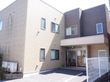 外観の様子である。外観は、ブラウンとベージュのツートンカラーである。出入口は自動ドである。建物前には、花や木が植えられている。