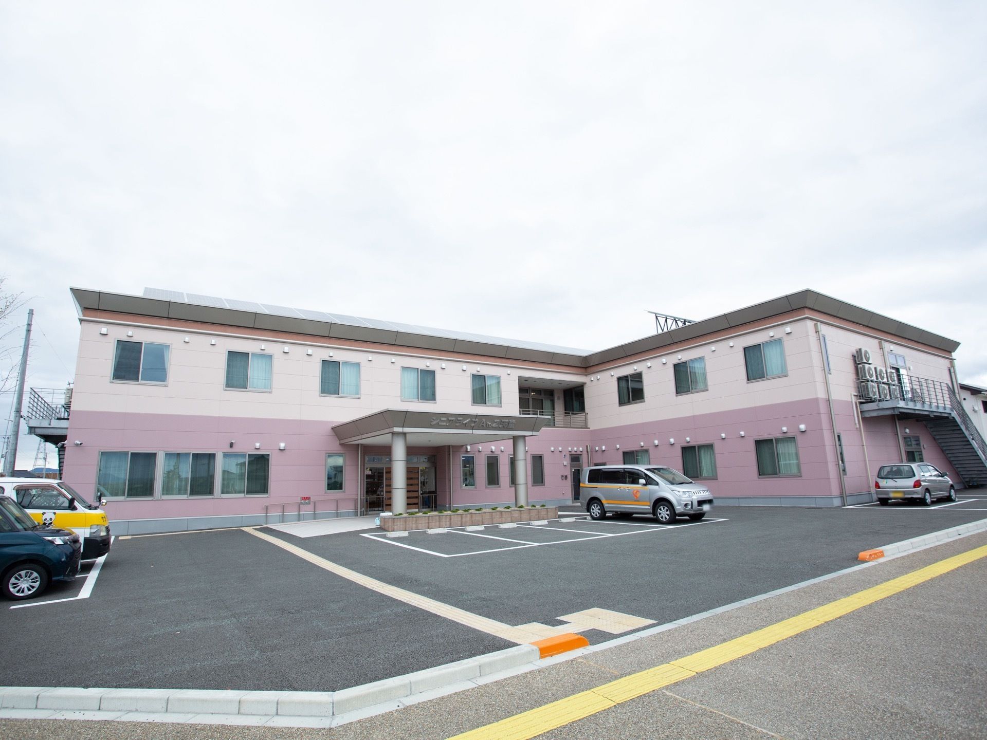 アスファルトで舗装された敷地に、Ｌ字型の２階建ての施設が建っている。白線で区画されたスペースに車が数台停められている。