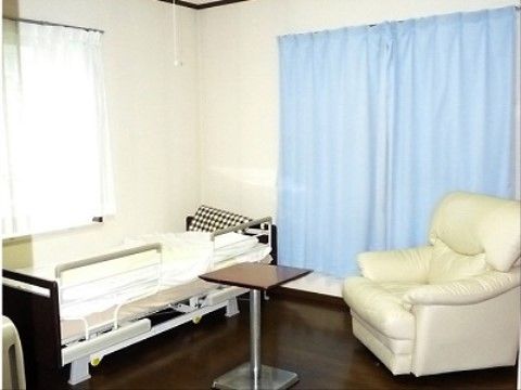 ゆったりとした居室の正面と横側に窓がある。壁際に介護用ベッドを配置し、傍に小さめの正方形のテーブルと白の１人掛けのソファを備えている。