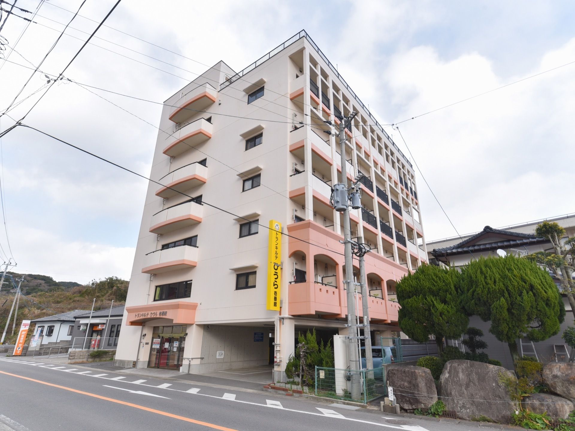 白とピンク色の外壁でかわいらしい雰囲気の建物になっている。施設の隣には郵便局があり便利である。黄色い施設名の看板がある。