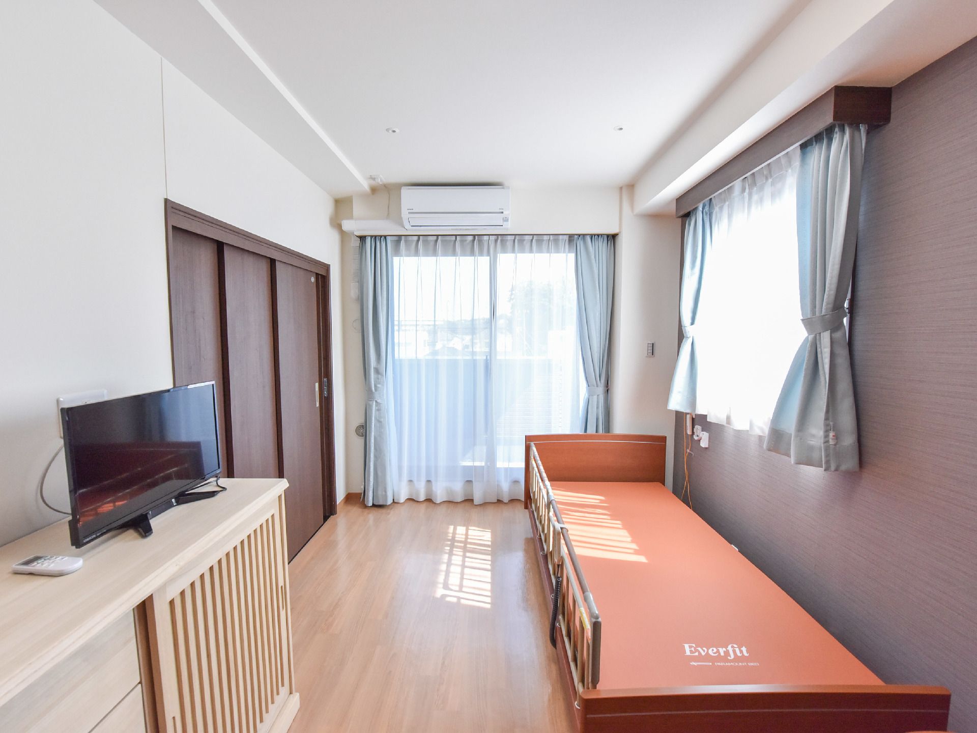 ウッドフロアの部屋で、壁際に電動ベッドがおいてある。ローボードの上にテレビが置いてある。ベランダ窓がある。