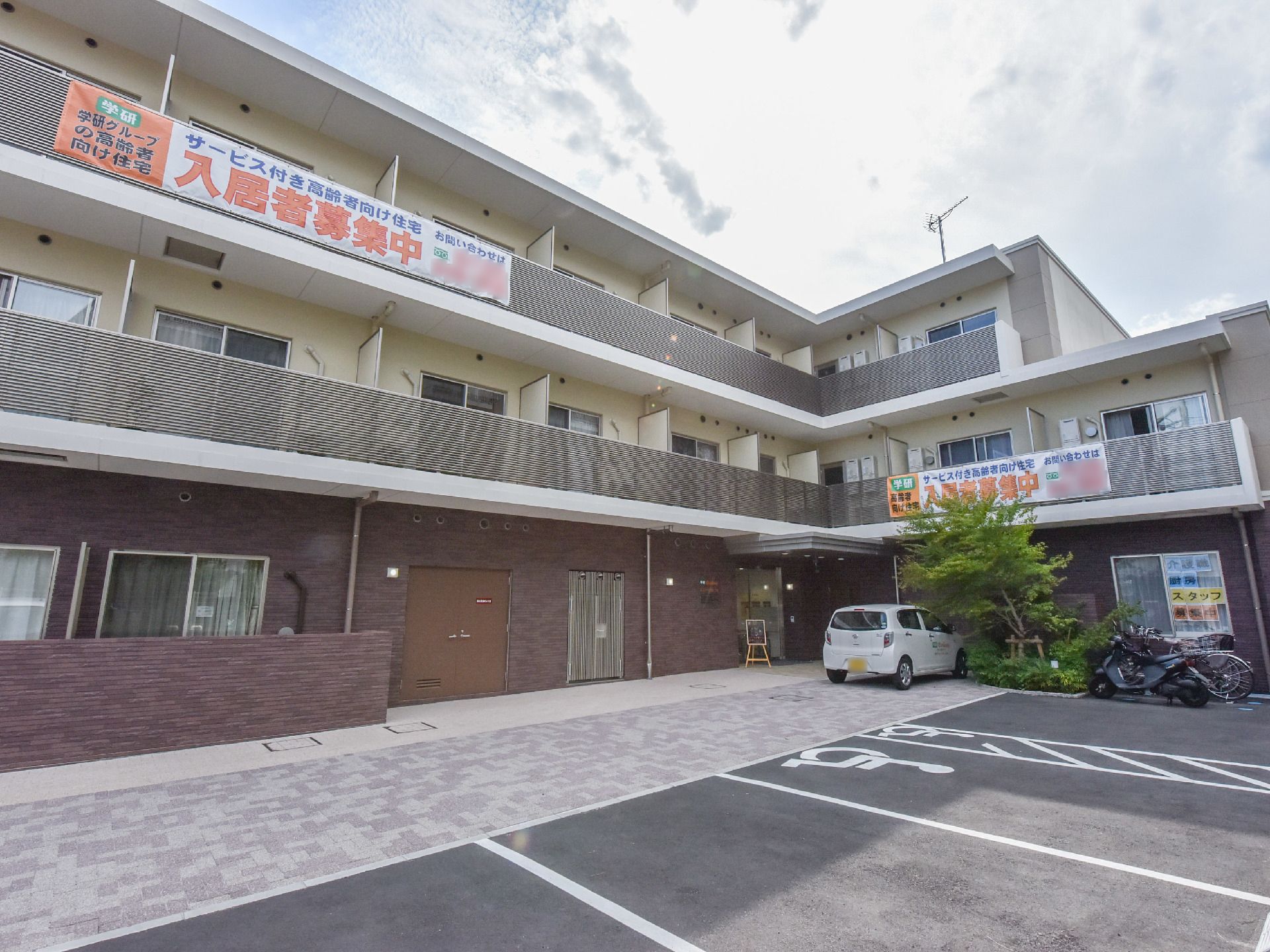施設の建物前の床はベージュ系のタイル敷きである。前面はアスファルト舗装の駐車場で、玄関側には車椅子マークがある。