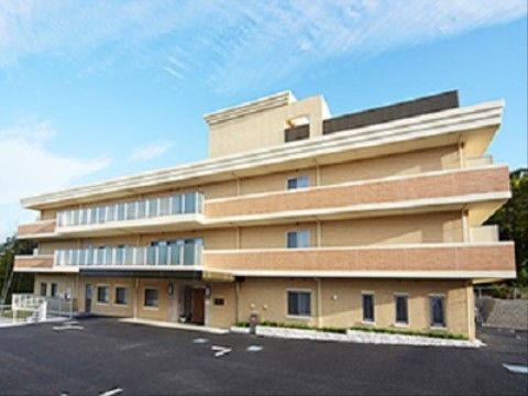 ３階建てのベージュを基調としたグループホームで、２階以上の部分にはベランダがある。施設前に駐車場を設けている。