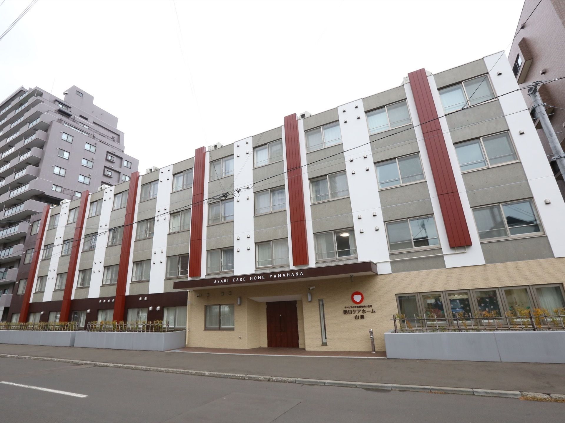 施設建物の正面玄関は目の前の道路に面している。木目調のドアはひさしの下の、やや奥まったところに配置されている。