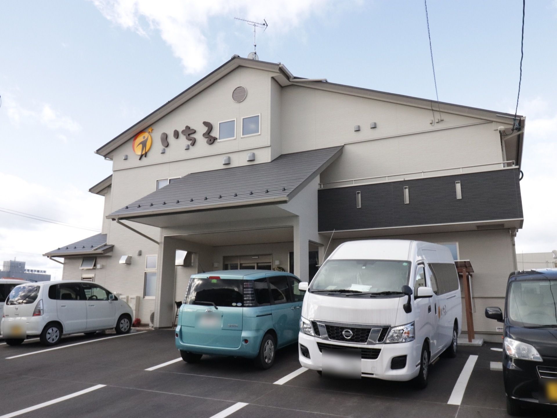 建物の上部の薄いグレイ色の壁に、黄色い円の中に人の姿を現した施設のロゴと、その隣に、茶色のひらがな文字で施設名を表示している。