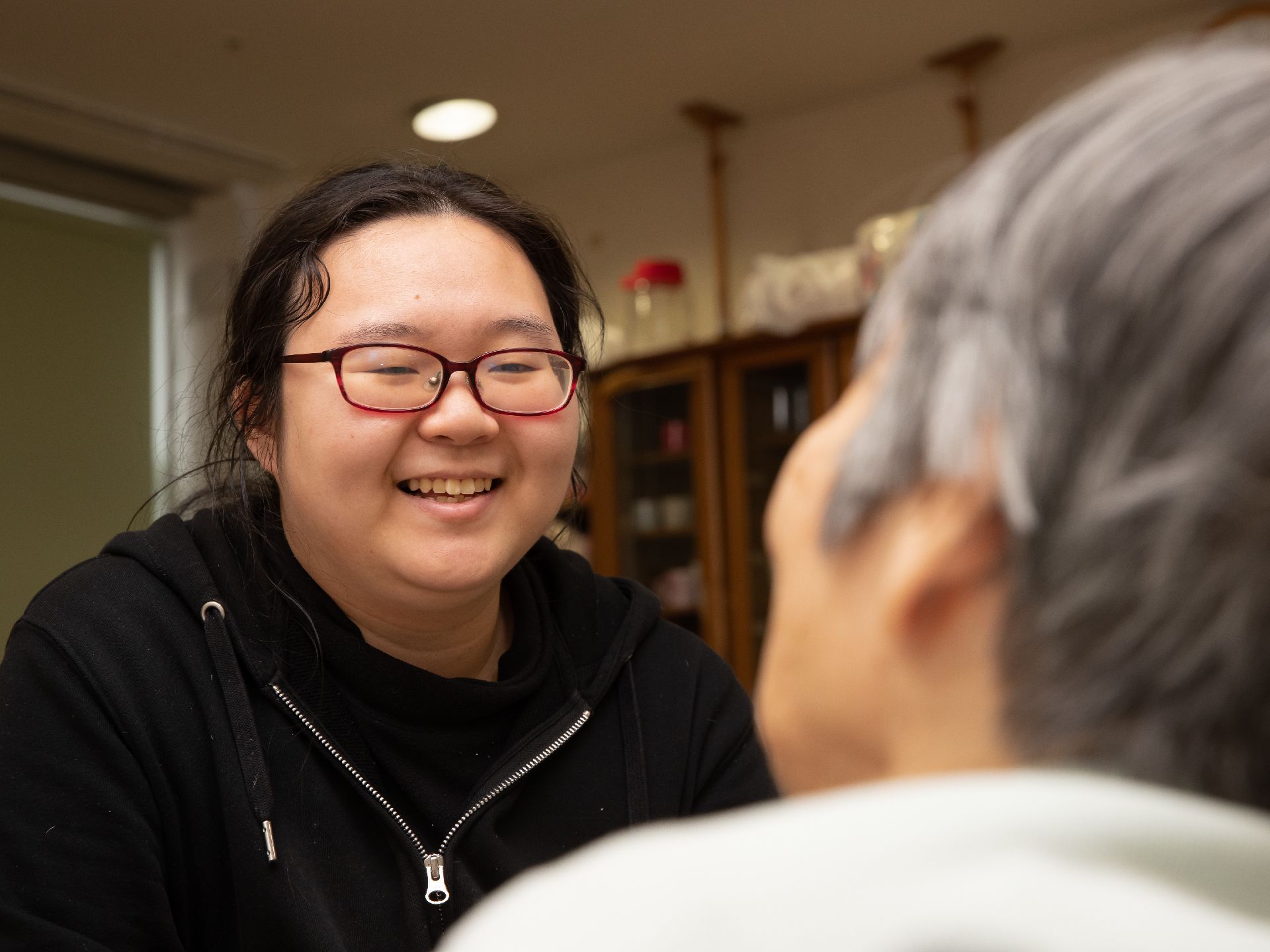 少しふっくらした女性スタッフが、入居者様と笑顔で会話している。後ろに見える棚は、しっかり地震対策がされている。
