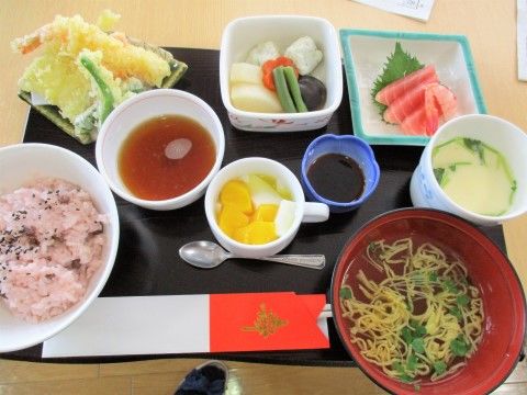 赤飯・刺し身・天ぷら・そばなど色とりどりの食事が、黒い盆の上に並んでいる。手前には袋に入った割り箸がある。