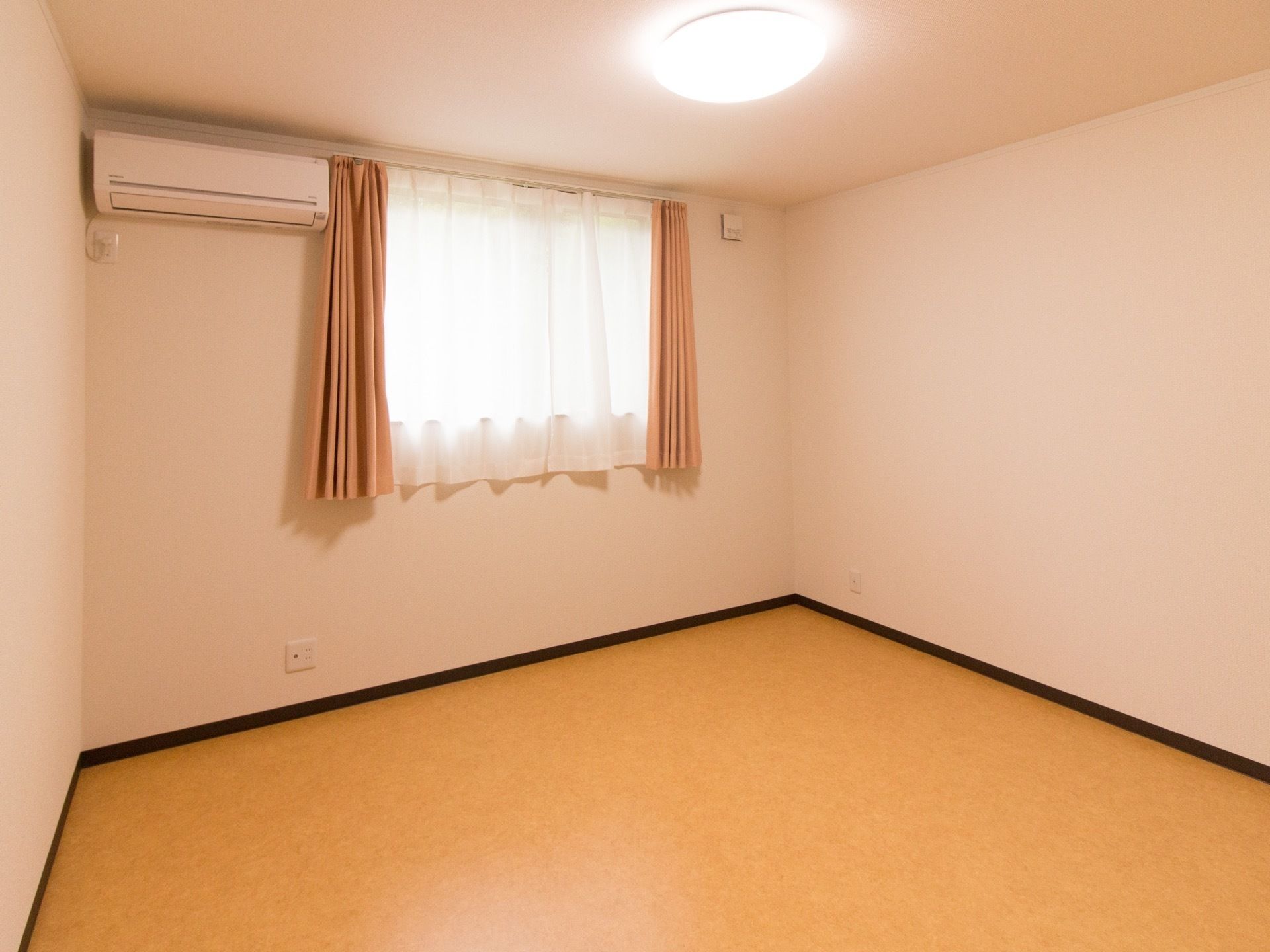 居室の奥には腰高窓があり、白いレースとベージュ色のカーテンが掛けてある。その左上に、エアコンが取り付けられている。