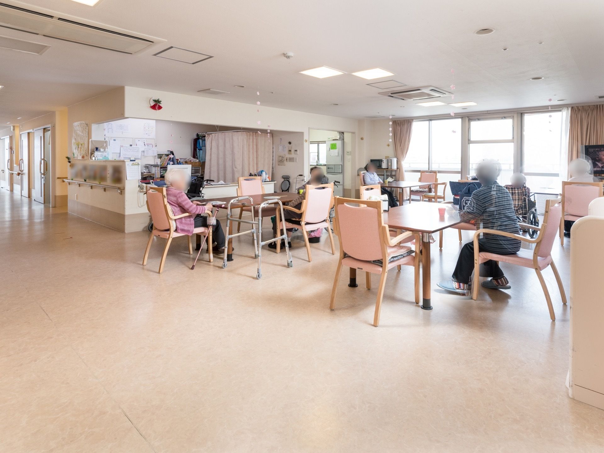 大きな窓が並ぶ明るい食堂。入居者様が集まり、テレビを見たり歓談をして過ごしている。車椅子や歩行器をご利用の方も移動しやすい広さがある。