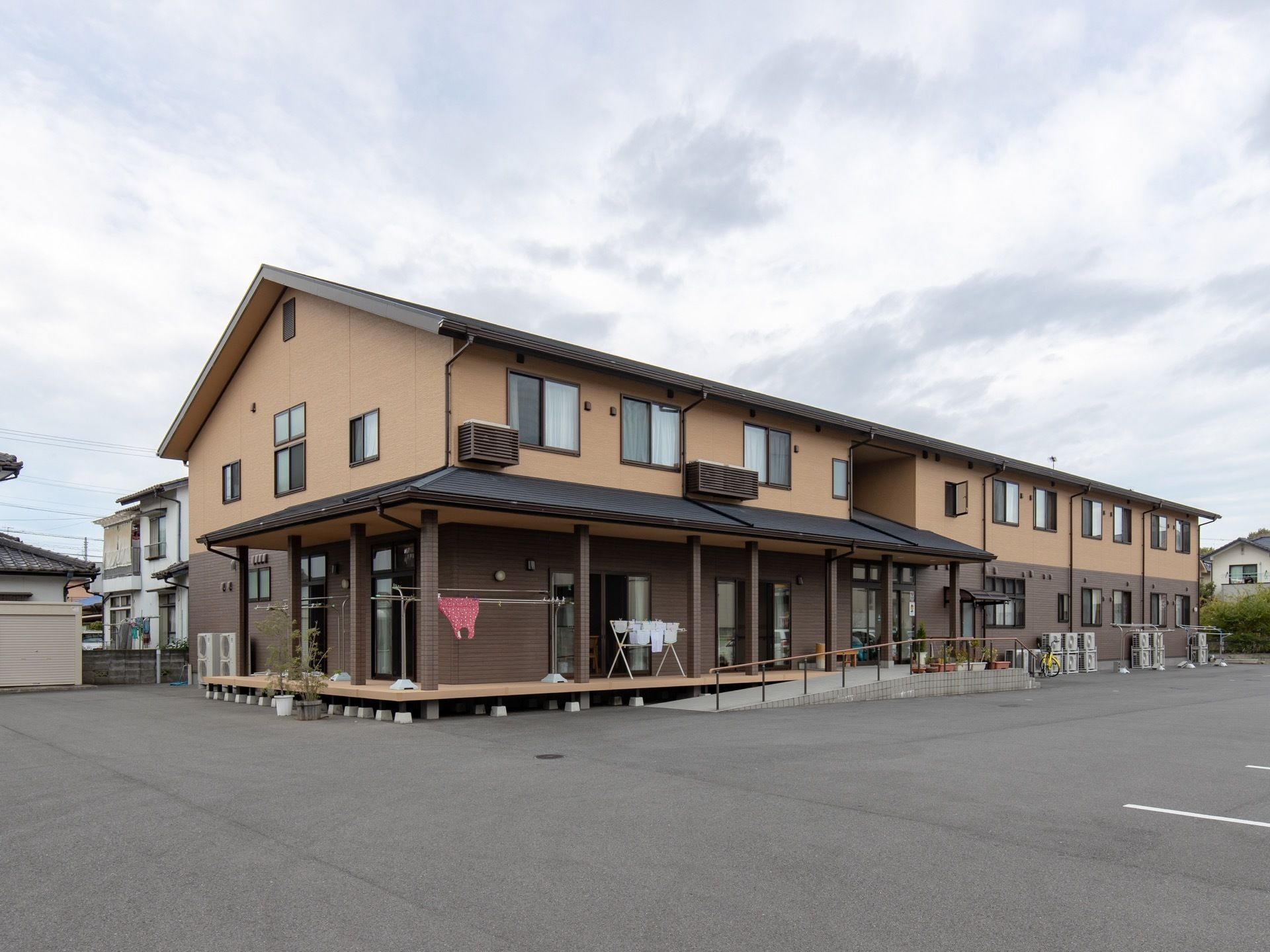 １階がこげ茶色で、２階が茶色い外壁の２階建ての建物がある。１階の縁側に洗濯物が干してあり、スロープが設置されている。