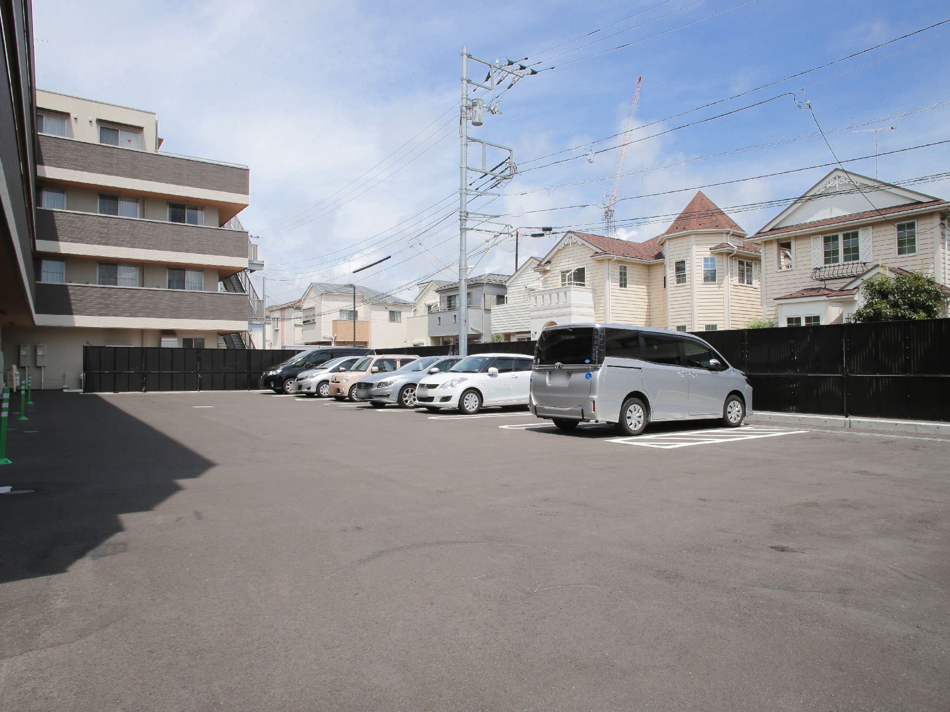 広々とした駐車場には、グレーのワゴン車やベージュ色の軽自動車など、数台の車が置かれている。その横には、一軒家が並んでいる。
