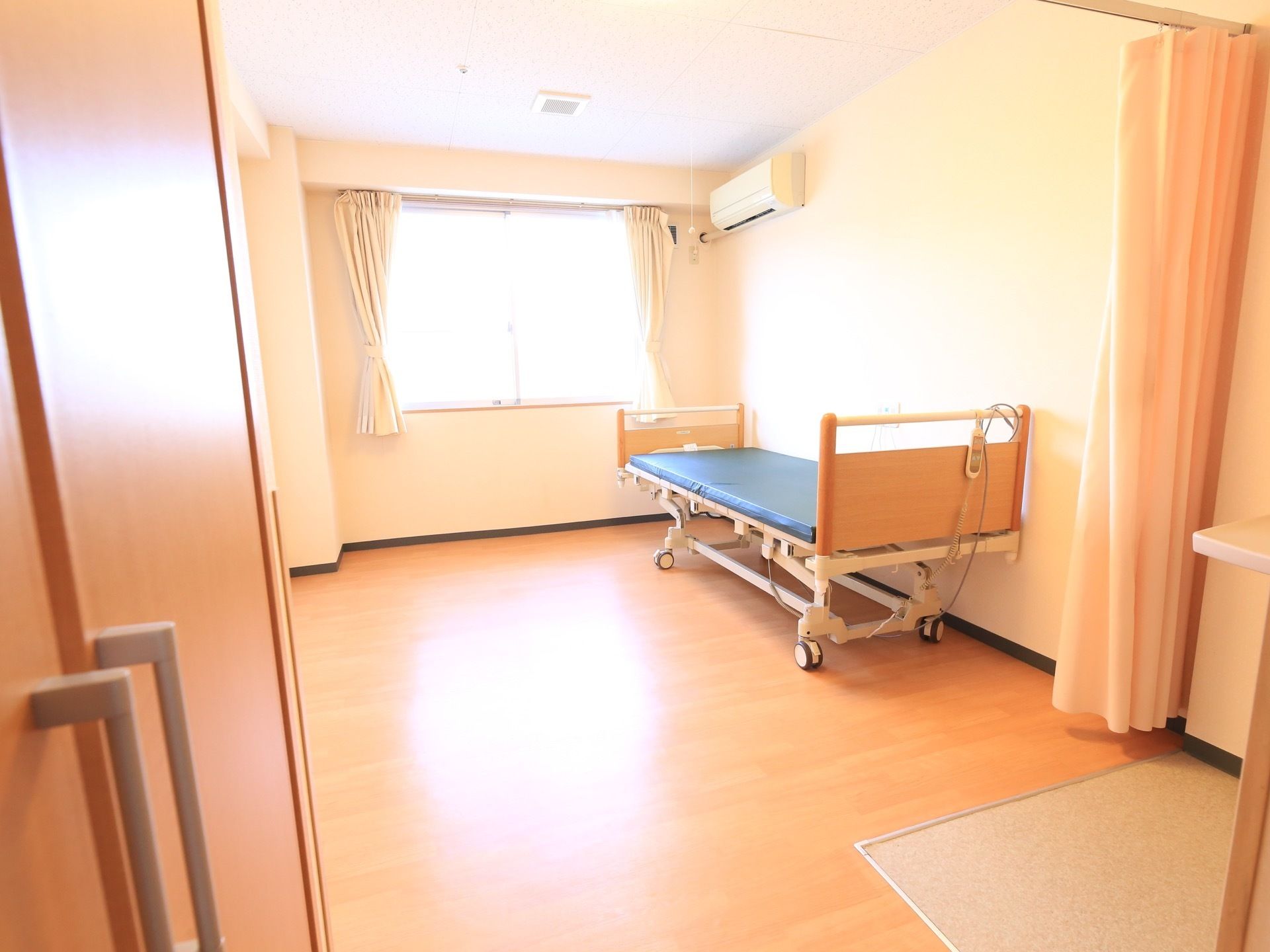 白を基調とした開放感のある明るい居室である。カーテンや介護用ベッド、エアコンが完備されている。広さが十分にある。
