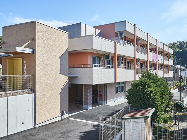 静かな住宅街にある3階建ての綺麗な建物。隣にエレベーターがある。周辺には樹々も見られ、落ち着いた雰囲気となっている。