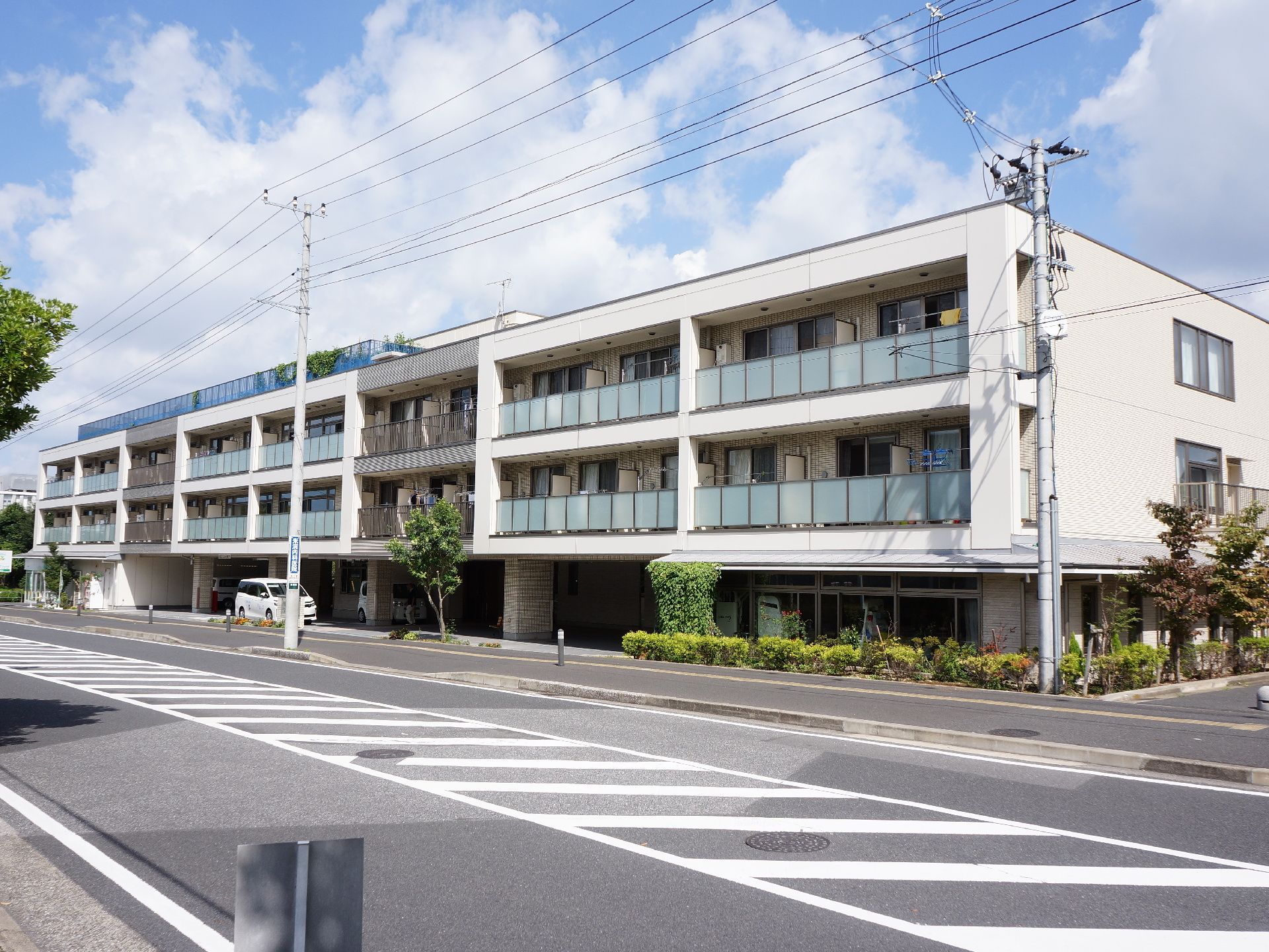 3階建ての施設で1階中央部に駐車場がある。2、3階にはベランダがあり、屋上には柵がしてある。外壁は白と濃いグレーになっている。