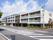 3階建ての施設で1階中央部に駐車場がある。2、3階にはベランダがあり、屋上には柵がしてある。外壁は白と濃いグレーになっている。