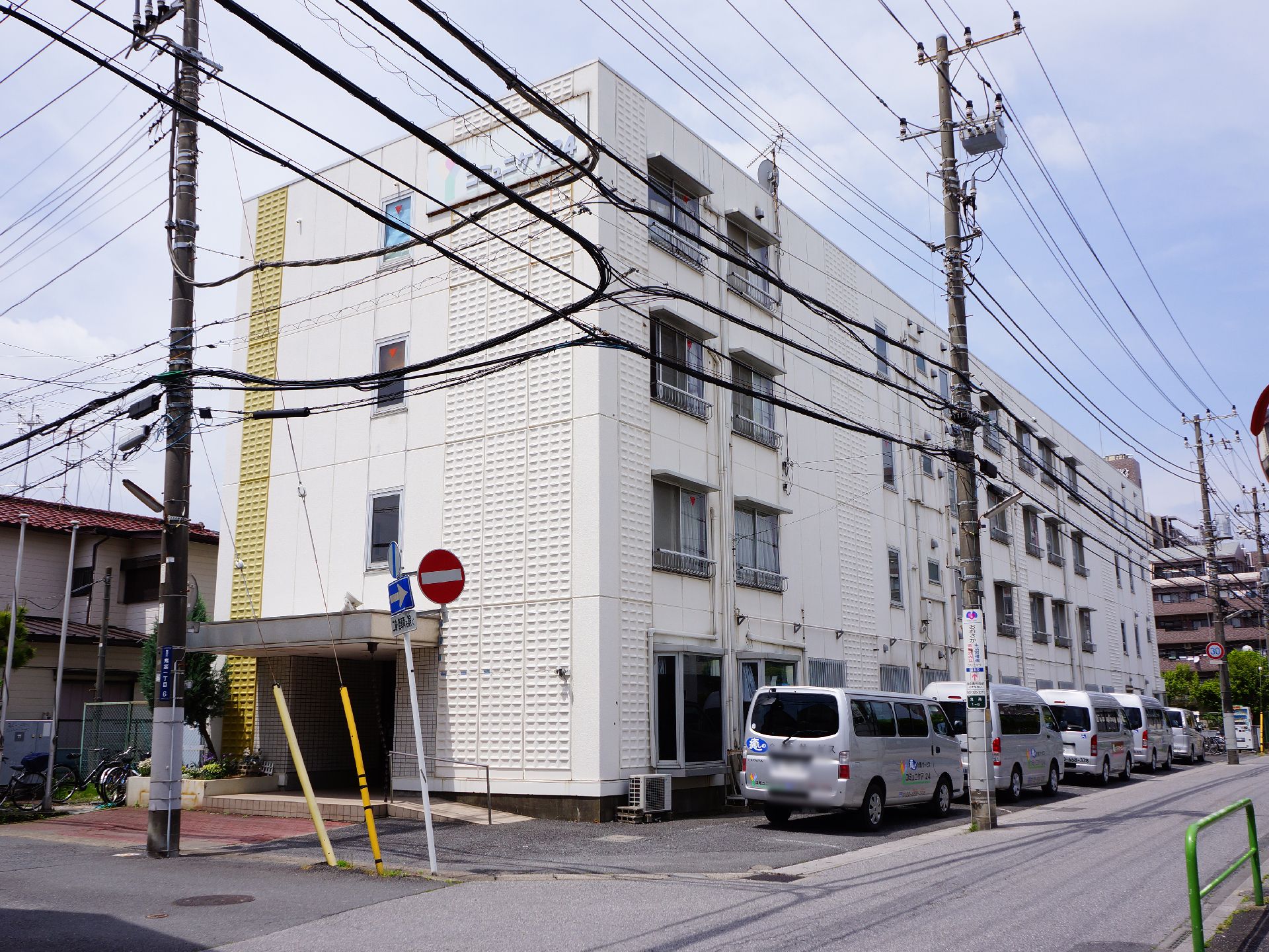 白い4階建の建物で手前を電柱と電線がはしっている。側面が入り口になっており角地で隣に車が何台も止まっている。