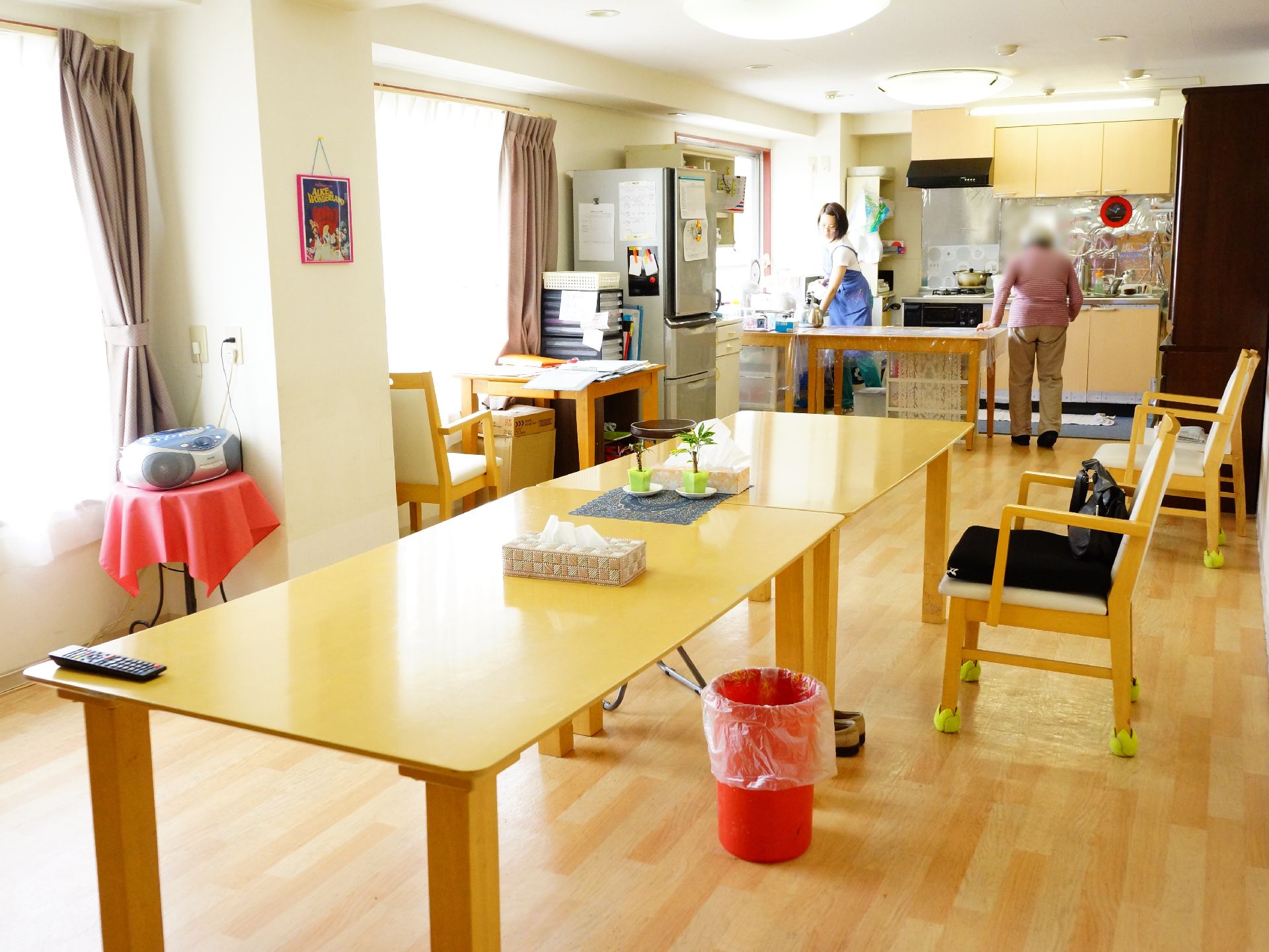 大きな窓から明るい光が屋内に差し込んでおり、奥には冷蔵庫やキッチンが見える。手前には大きな木目調のテーブルが並ぶ。