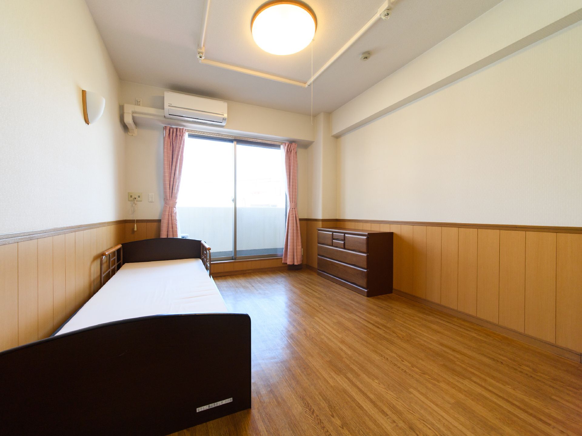 室内にチェスト、ベッド、エアコンが設置されている。窓の横、ベッドの頭側の壁にナースコールが付いている。