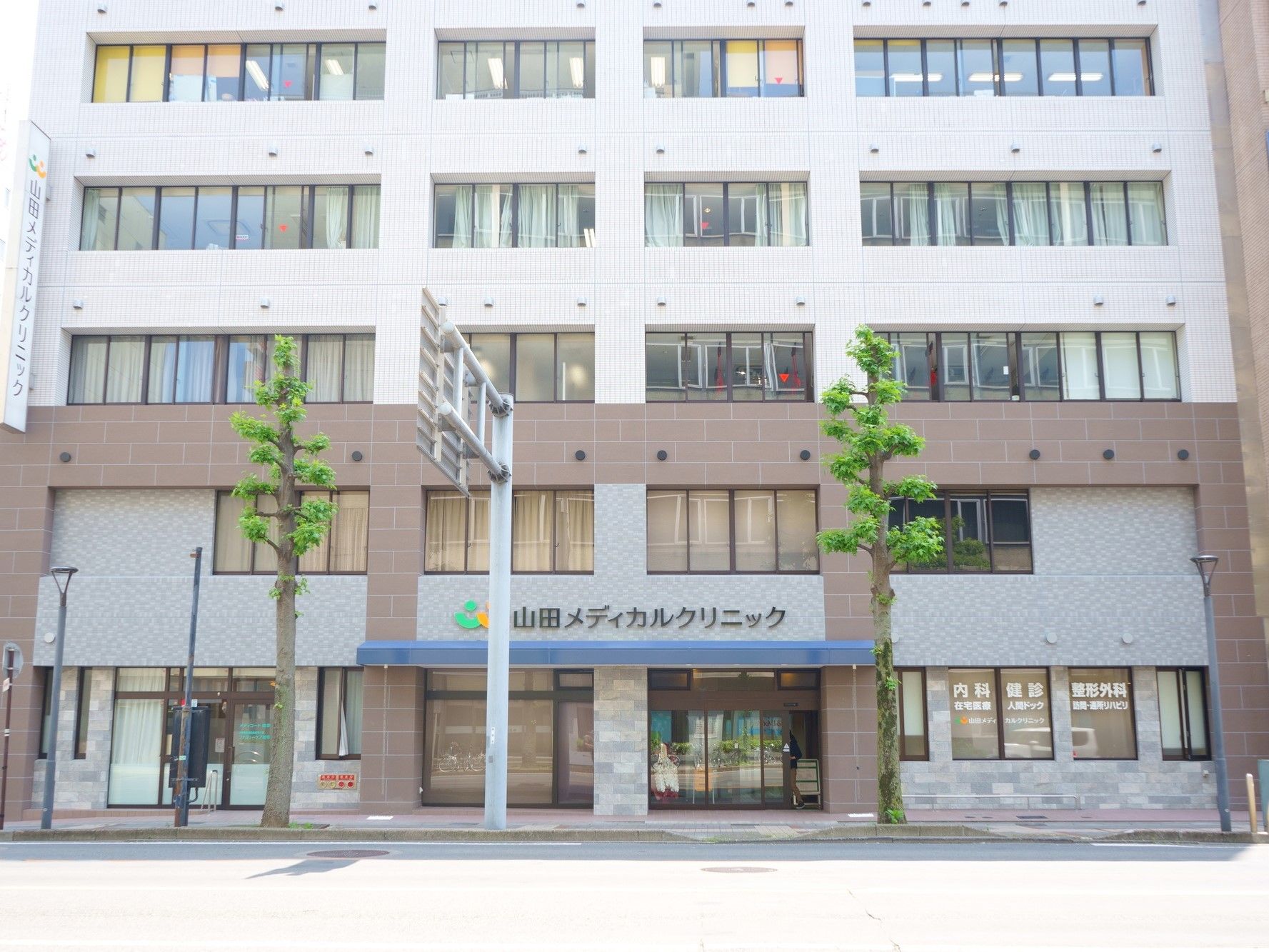 外壁は白のタイル張りである。ひさしと診療所の看板のグリーンがアクセントになっている。建物の前に街路樹が立っている。