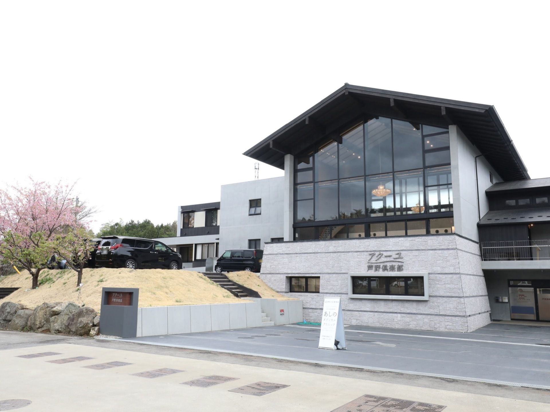 正面に窓ガラスが多数設置されている建物が建っている。その横の階段を上ると駐車スペースがあり、車が停まっている。