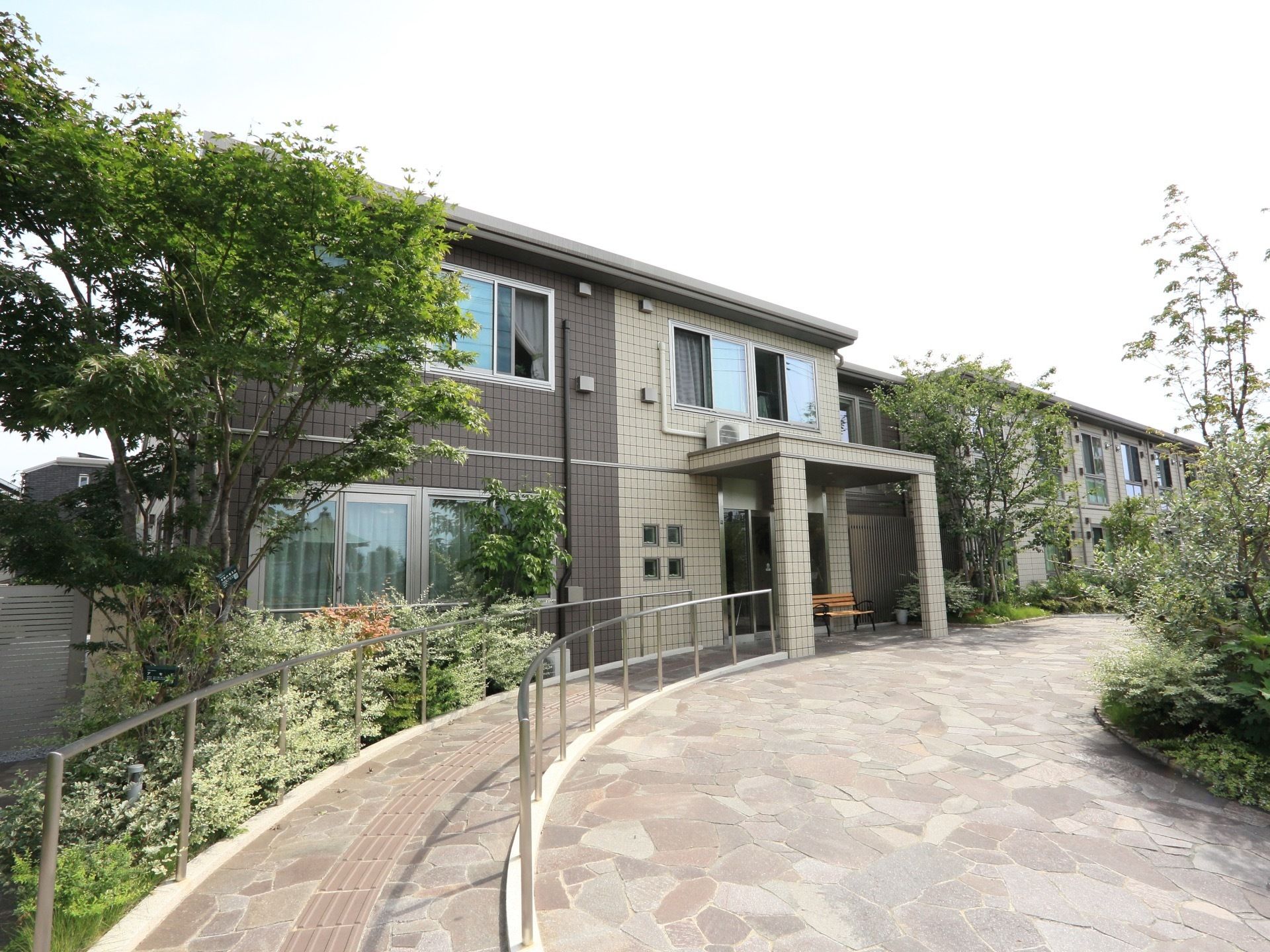 緑豊かな住宅街にたたずむ施設の様子。幅の広い歩道には手すりが備わっている。玄関の自動ドアの前にベンチが置かれている。