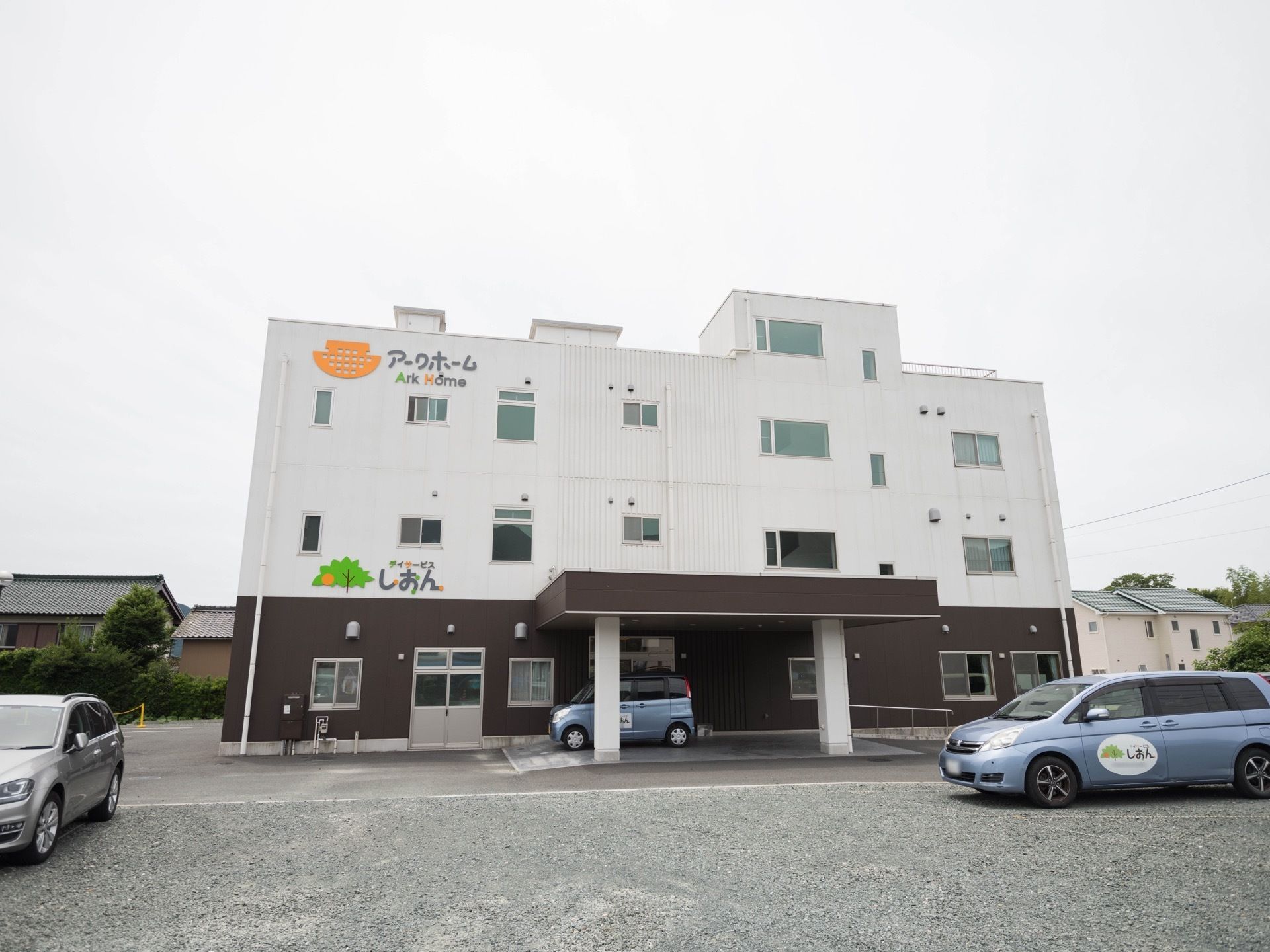 １階が茶色い、３階建ての白い建物が建っている。建物の背面に数件の民家があり、敷地内に数台の車が停められている。