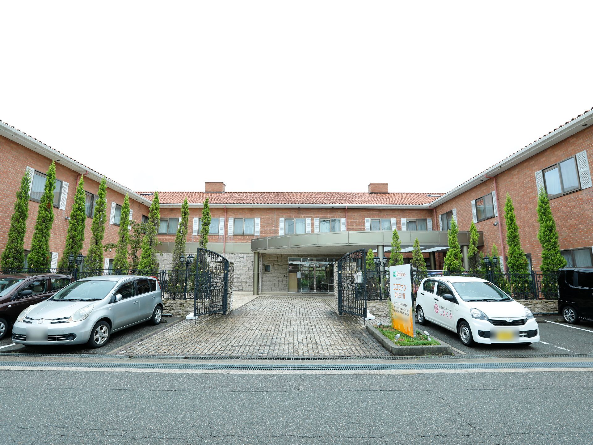 施設の前には駐車場が設けてある。フェンスと木々が植えてある。茶色の外壁の建物がある。正面玄関のゲートが開いた状態になっている。