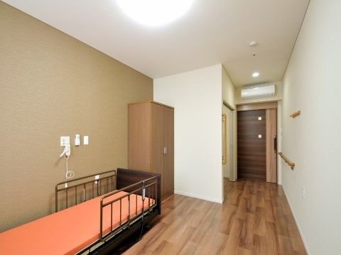 深みのある木目調の床を取り入れた居室は、落ち着いた雰囲気である。壁際に介護用ベッドを配置し、横側に収納家具を備えている。