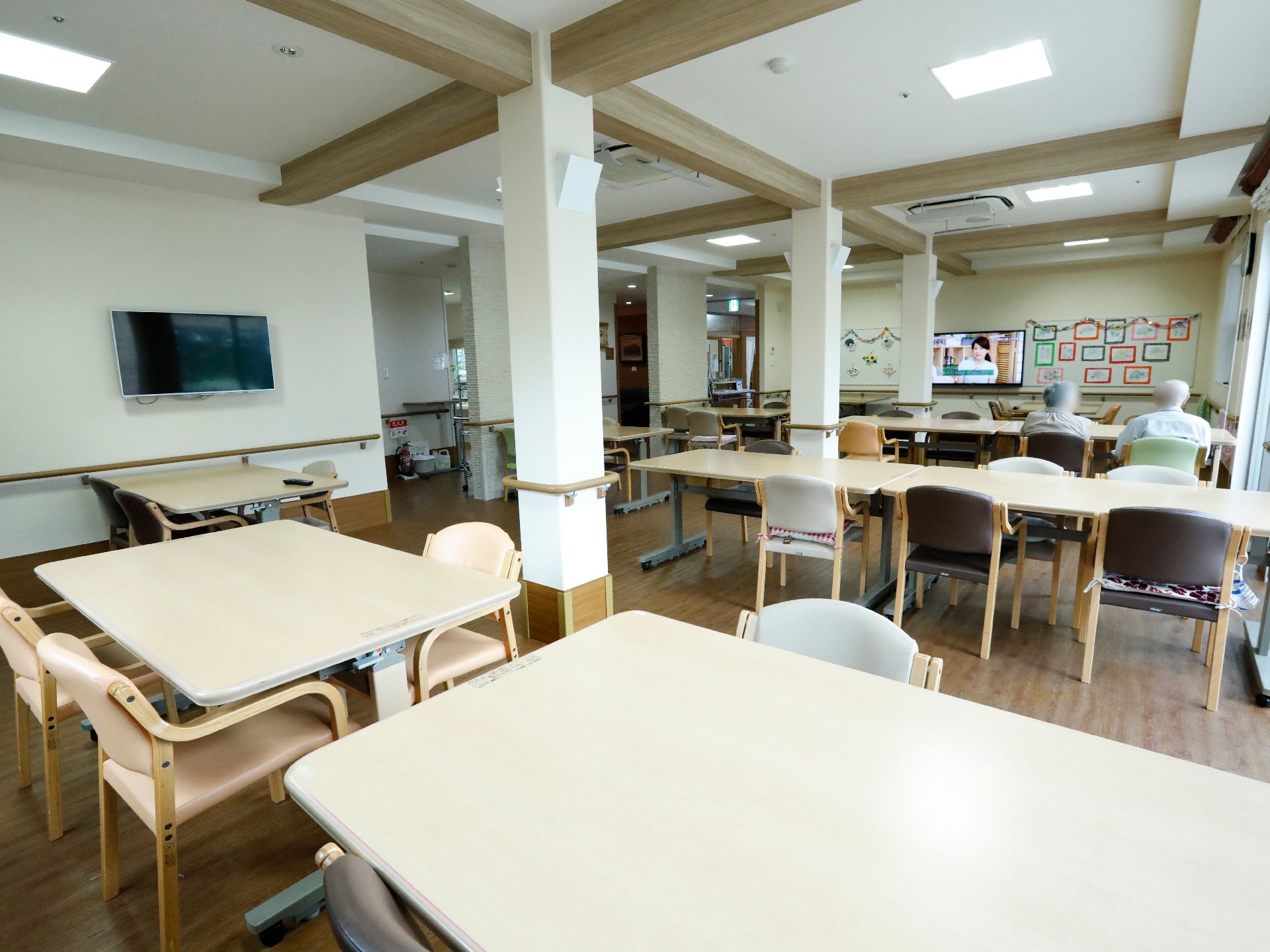 フローリングの食堂である。テレビがついていて、入居者様が二人テレビを見ている。壁にガーランドなどで飾り付けがしてある。