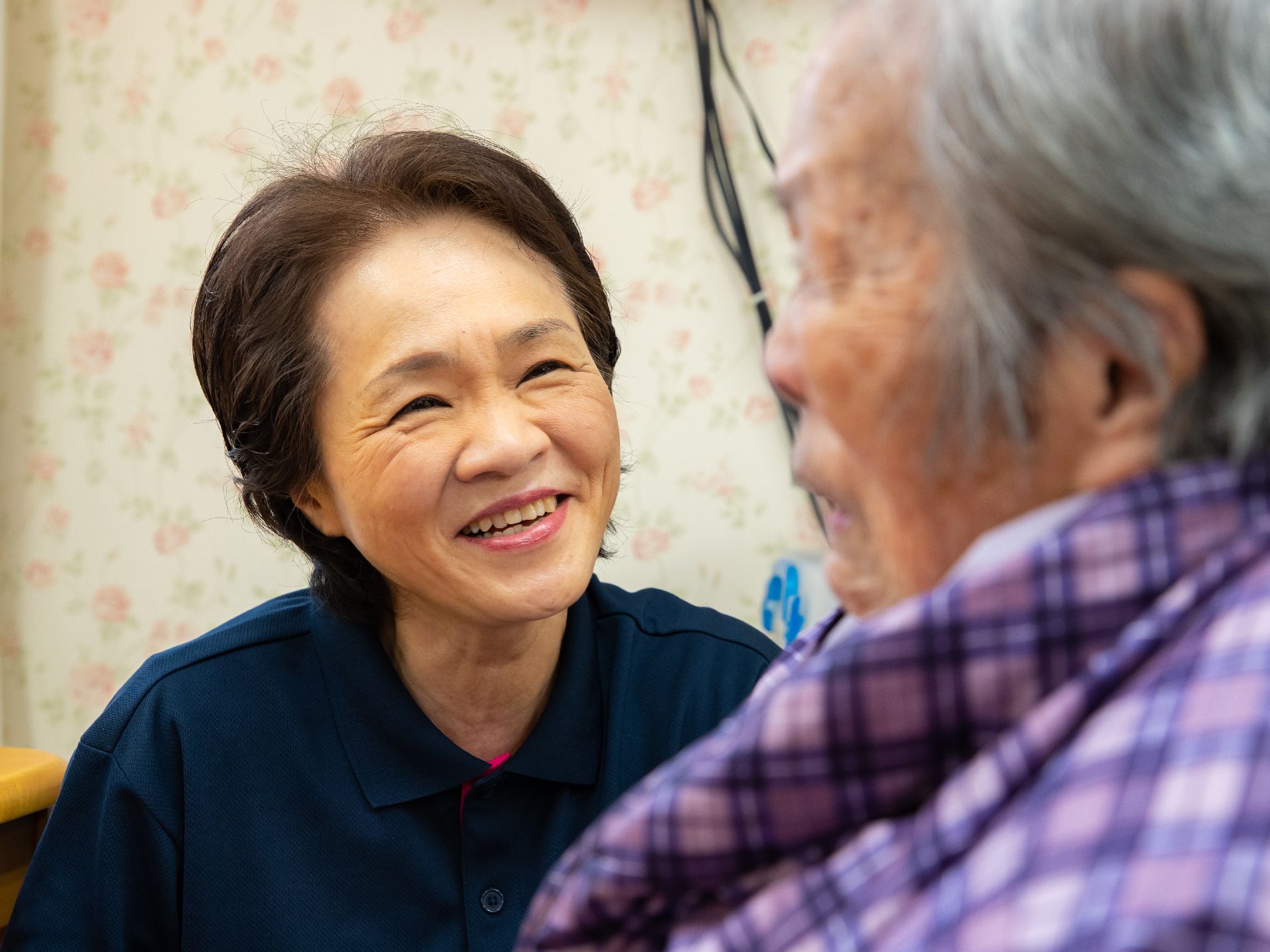 花柄の壁の前で、女性スタッフが座っている入居者様に目線を合わせて笑いかけている。左側には、テーブルがある。