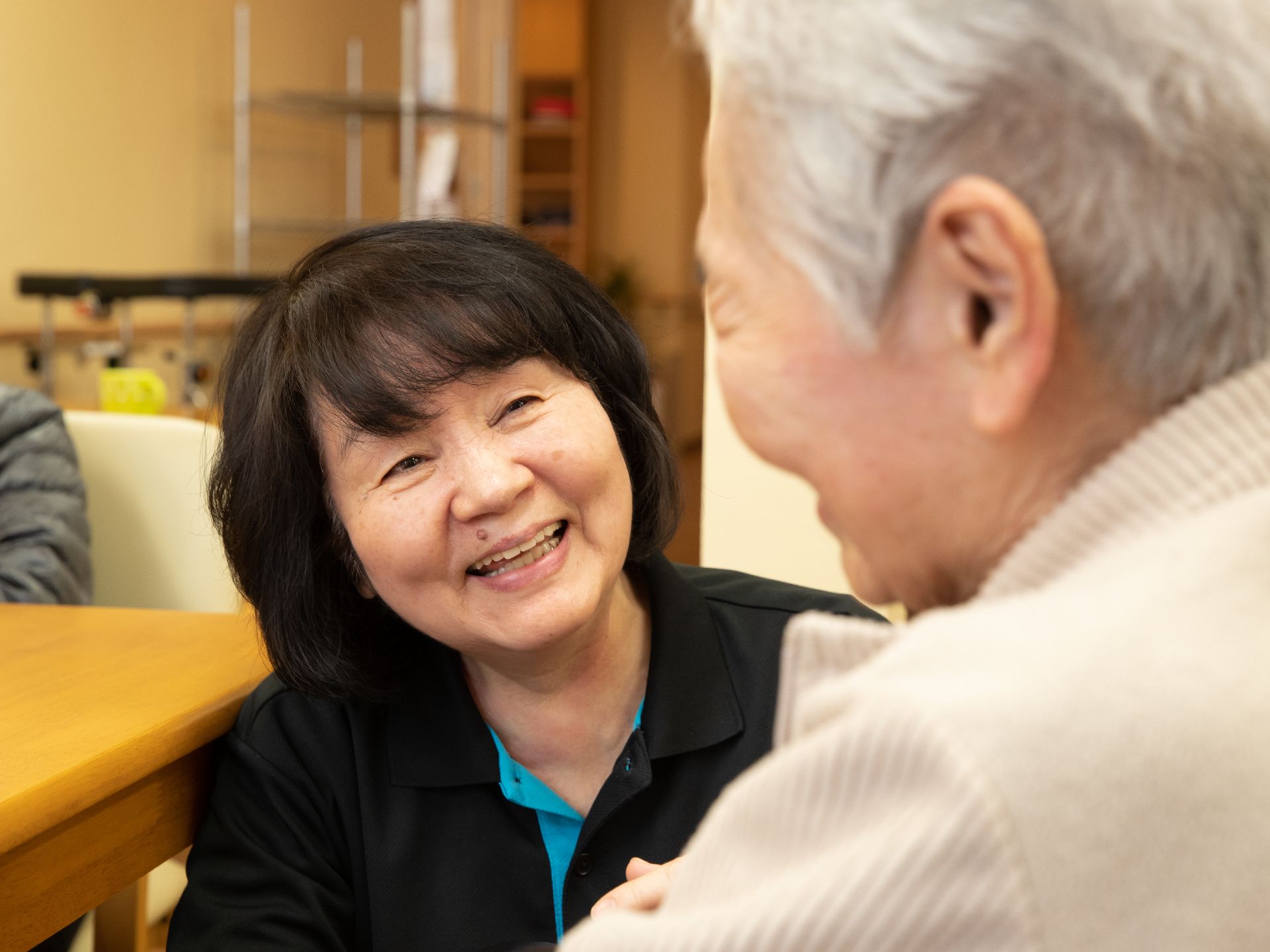 テーブル席に着いている入居者様と女性スタッフが微笑み合っている。左後ろの壁には、手すりが設置されている。